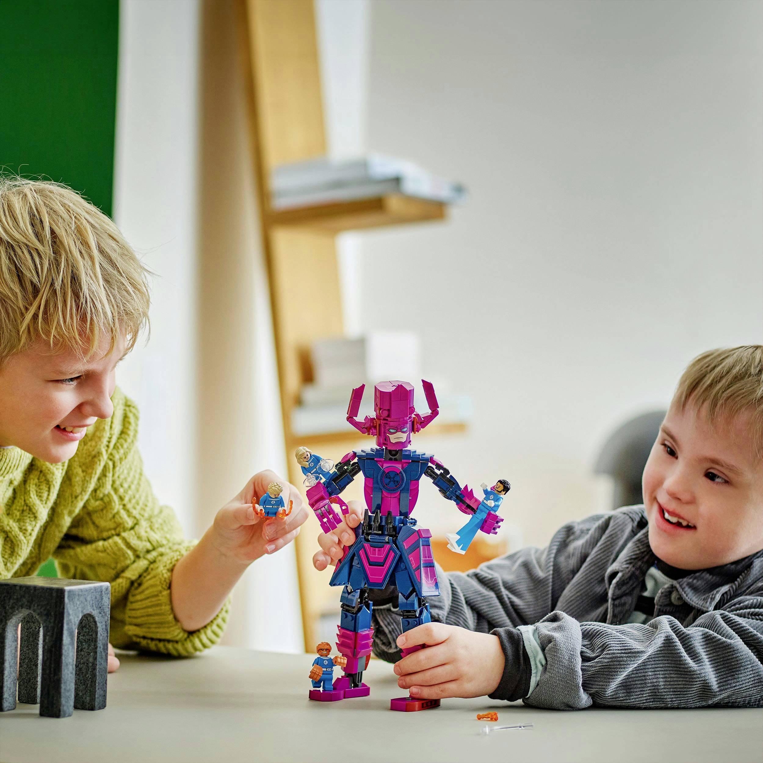 Two boys are playing with colorful figures and blocks on a table. One of the boys is holding a figure, the other is standing upright in the middle.