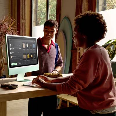 Two people are sitting at a table with a computer screen displaying various clothing items. They appear cheerful and are in a modern room with large windows.