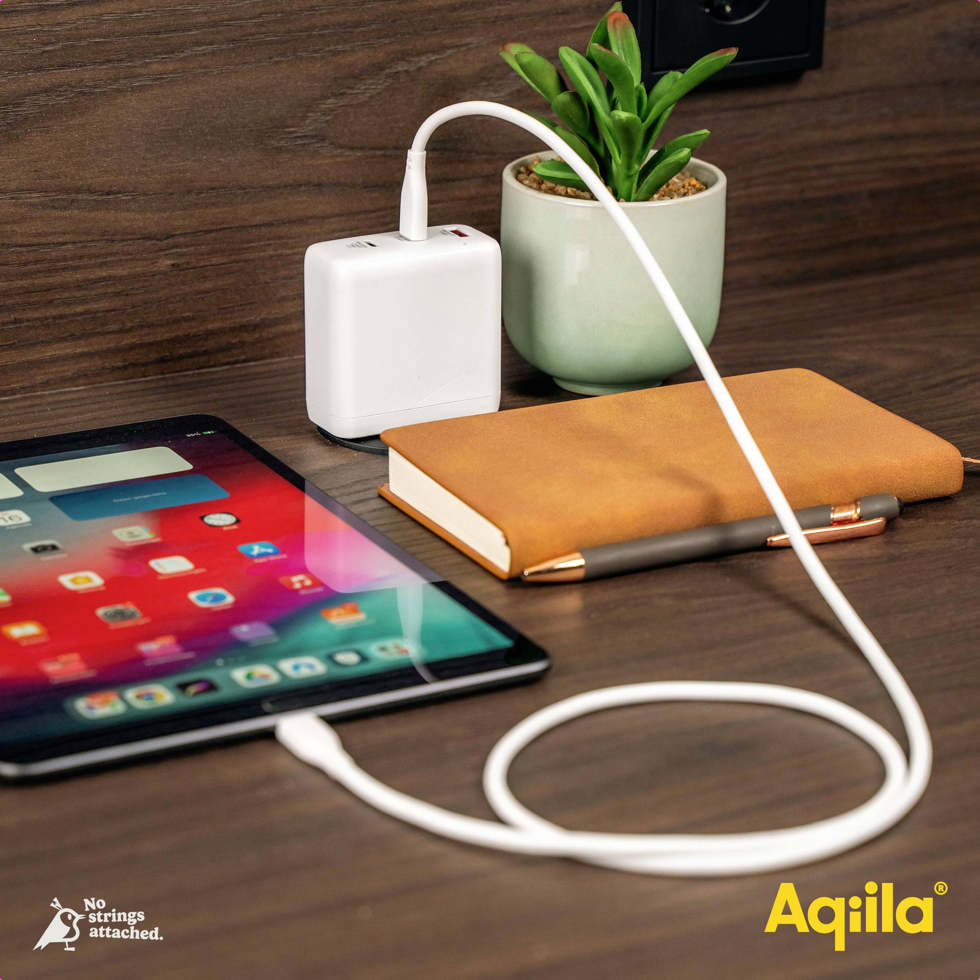A tablet is being charged via a charger on a wooden table. Next to it are a notebook, a pen, and a small plant.