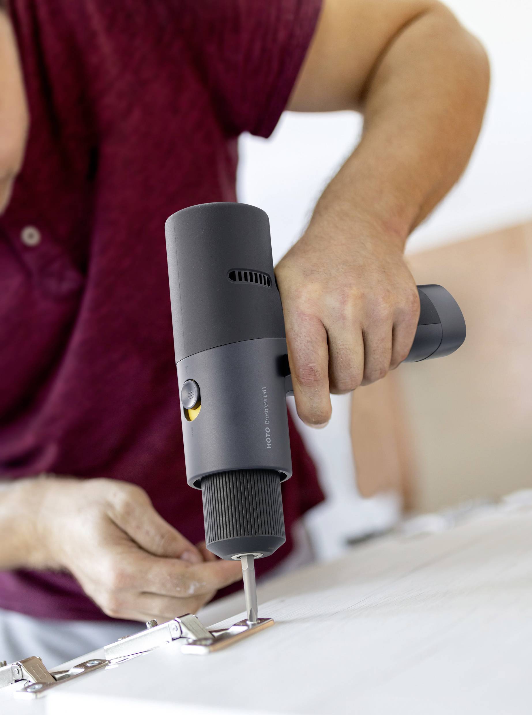 A person wearing a red shirt is using an electric screwdriver to turn a metal screw into a mounting bracket on a worktop.