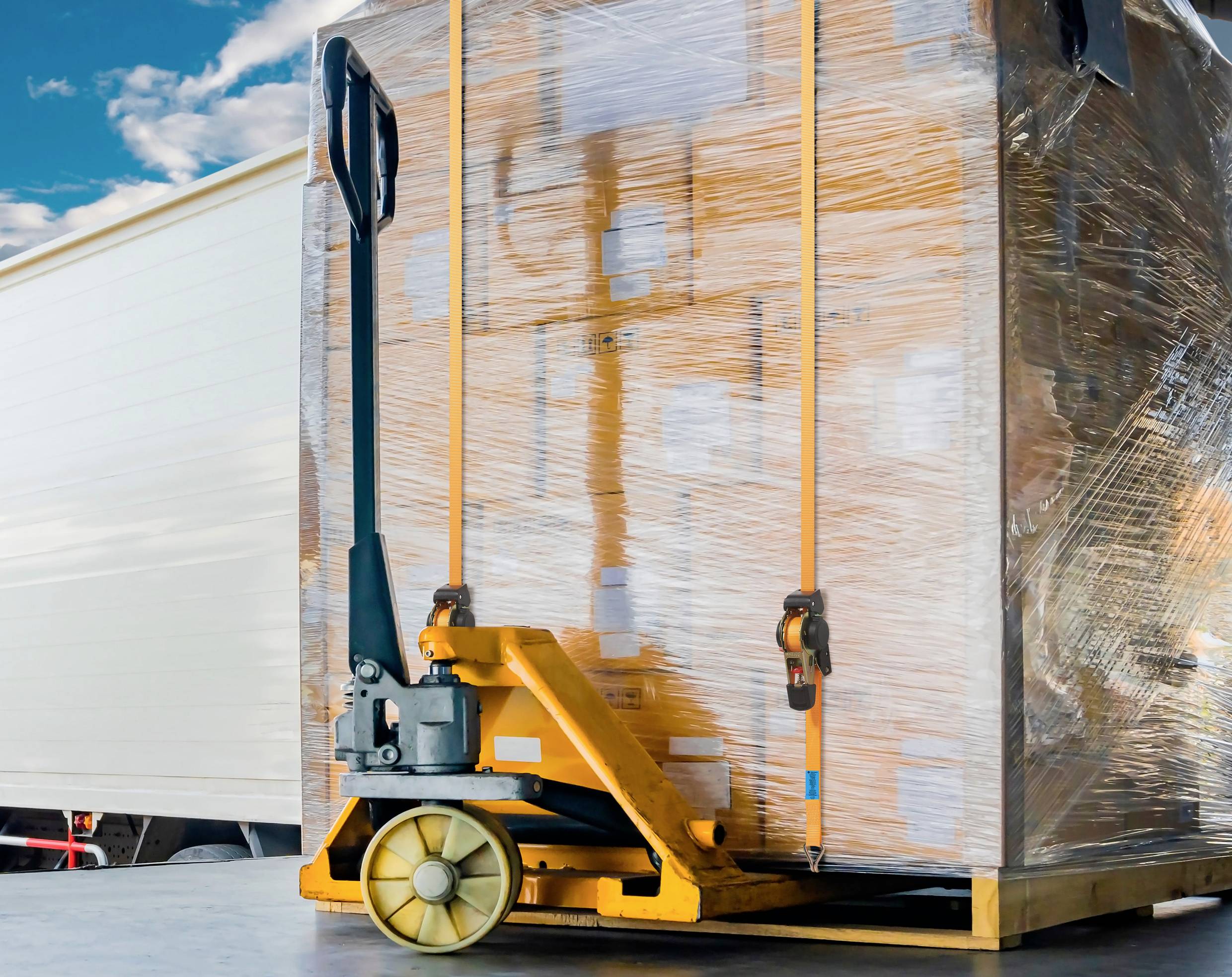 A hand pallet truck is transporting a pallet wrapped in stretch film with cardboard boxes in front of a lorry. In the background, the sky is visible.