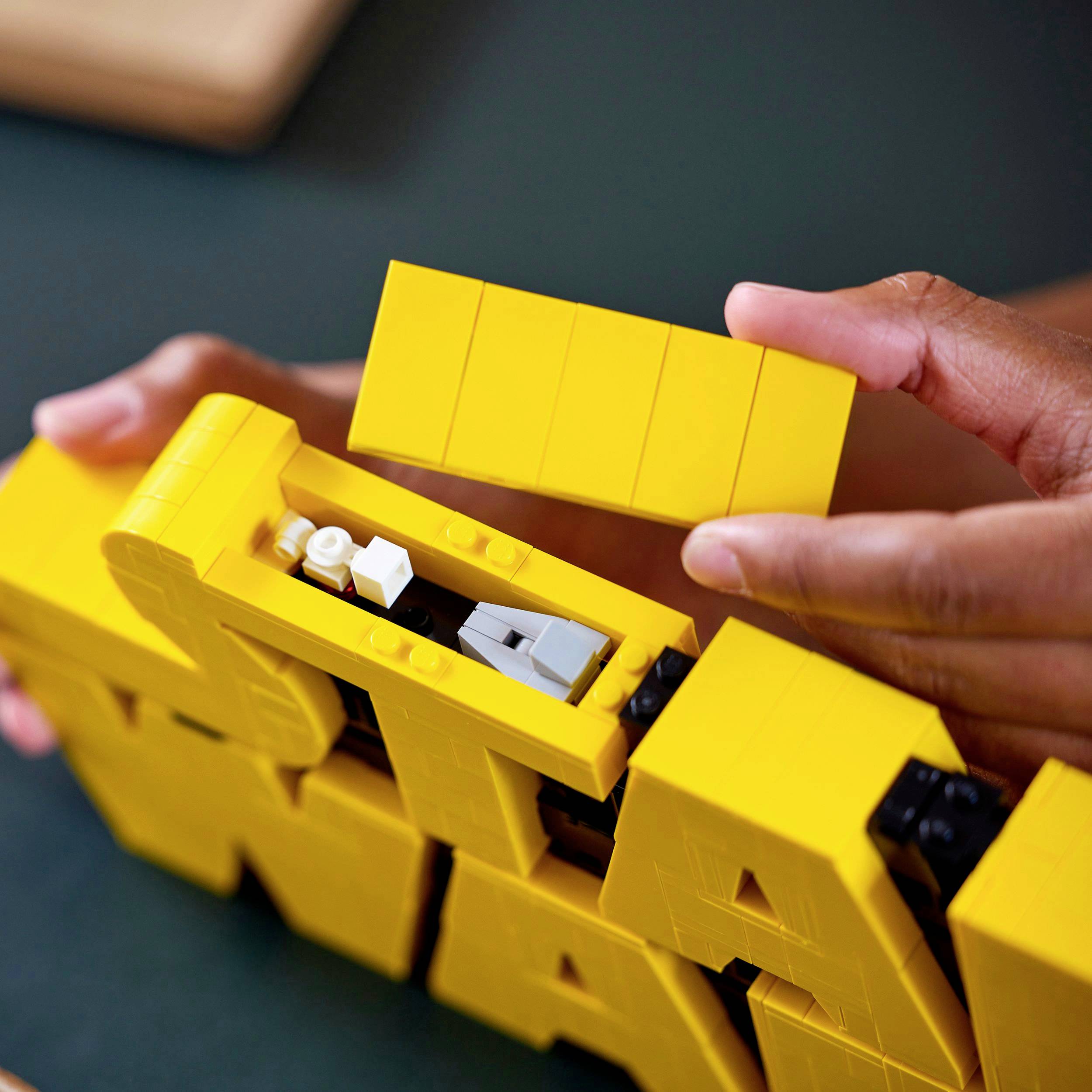 Hands lift a yellow LEGO building plate to reveal the hidden mechanism of a model underneath.