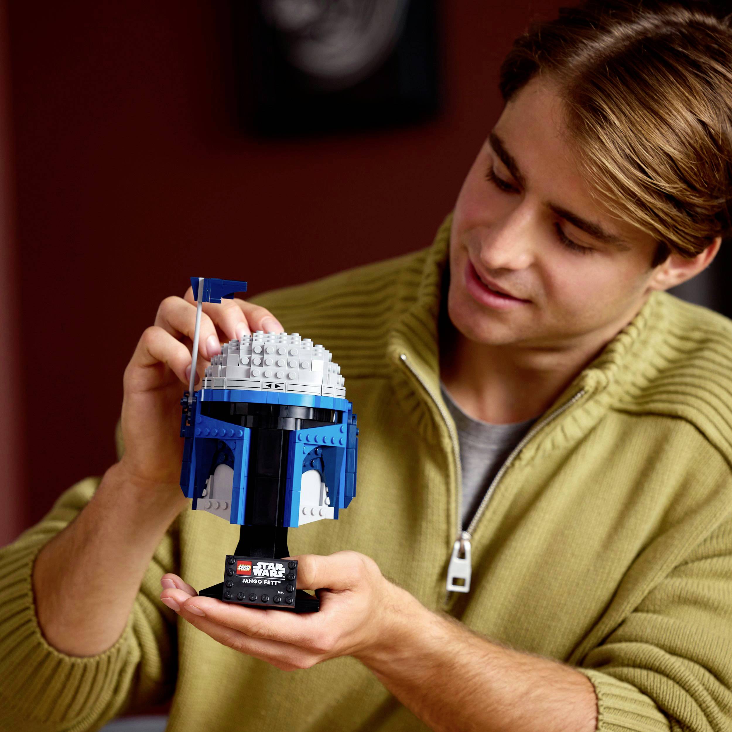 A man is holding a Star Wars LEGO helmet model. He is wearing an olive green jumper and studying the model carefully.
