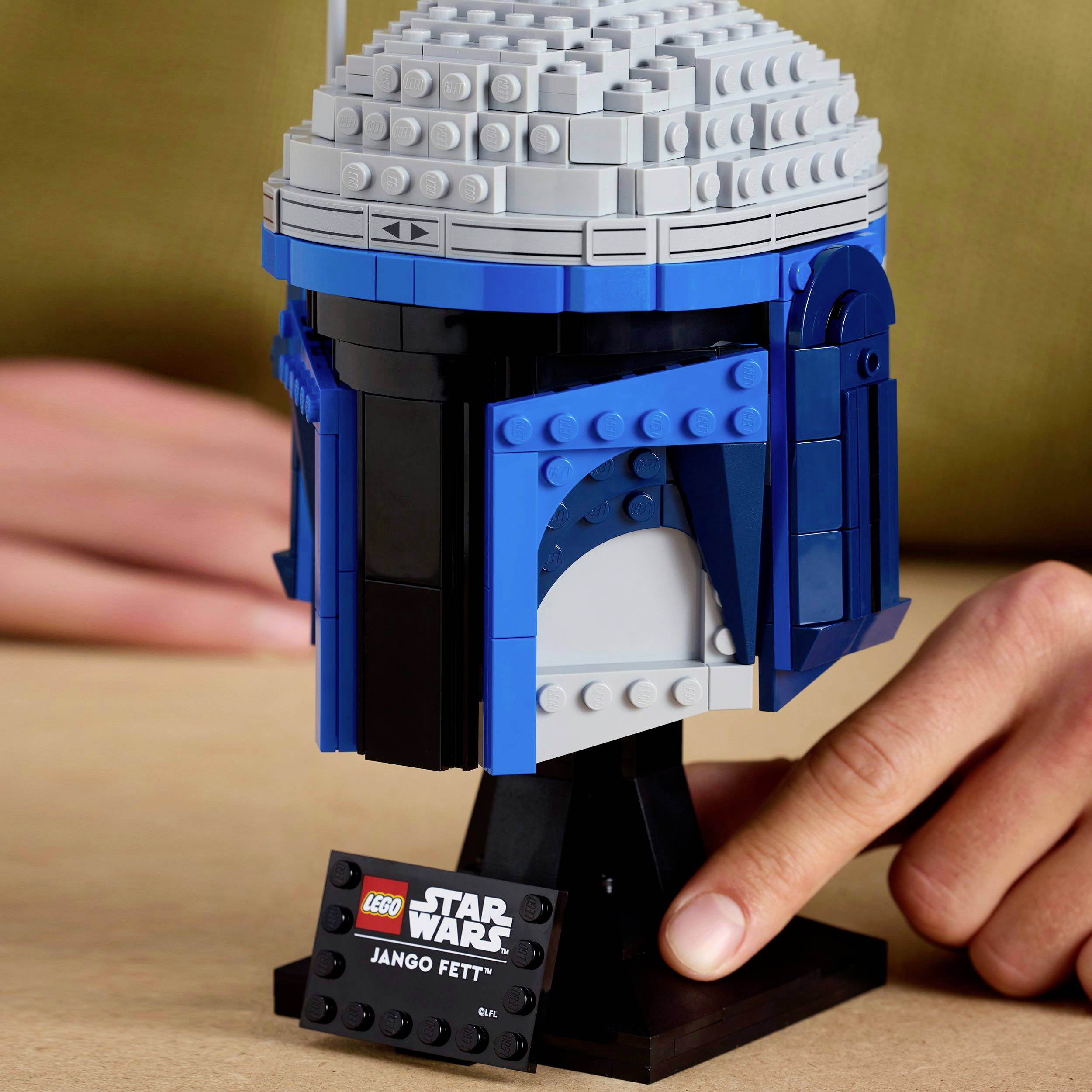 A LEGO Jango Fett helmet from Star Wars is displayed on a table. A hand is pointing at the black shield with the name.