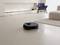 A black robotic vacuum cleaner is cleaning the floor in a modern living room with a fireplace, a blue armchair, and a rug.