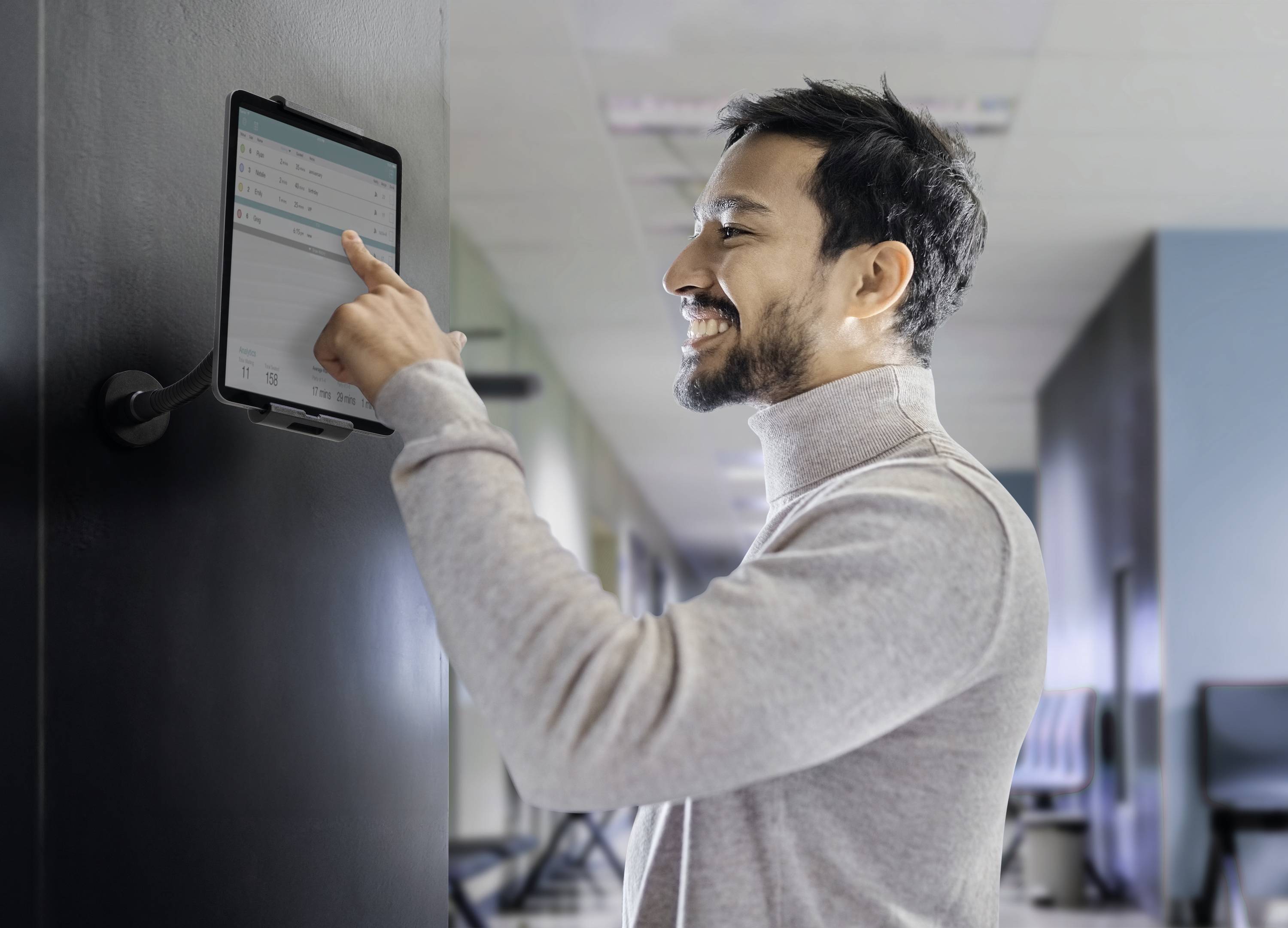 A man is standing in front of a tablet mounted on a wall, smiling and tapping on it. He is wearing a polo neck jumper. The room is decorated in a modern style.