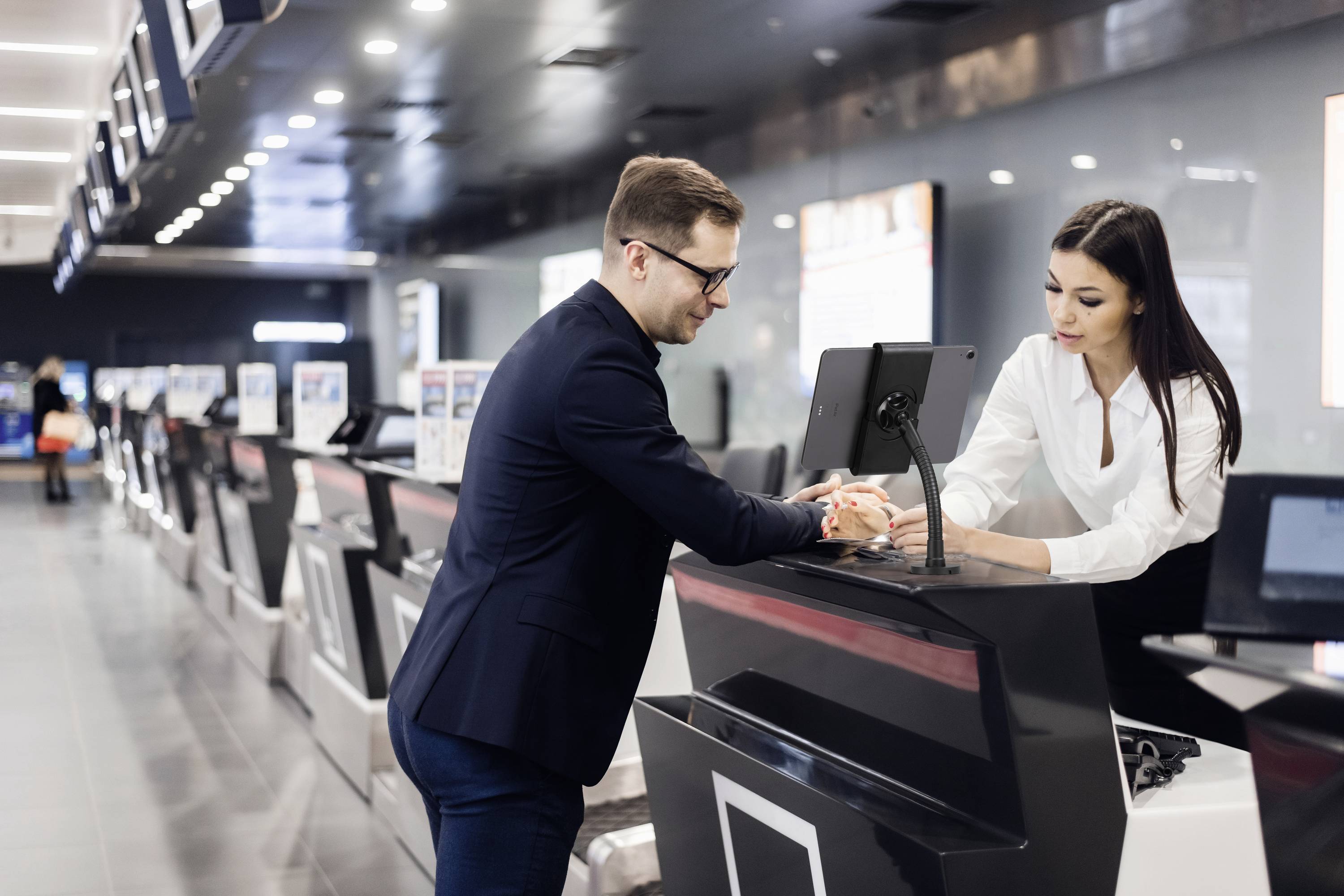 A man and a woman are standing at an airport check-in desk. The man hands documents to the woman while she assists him.