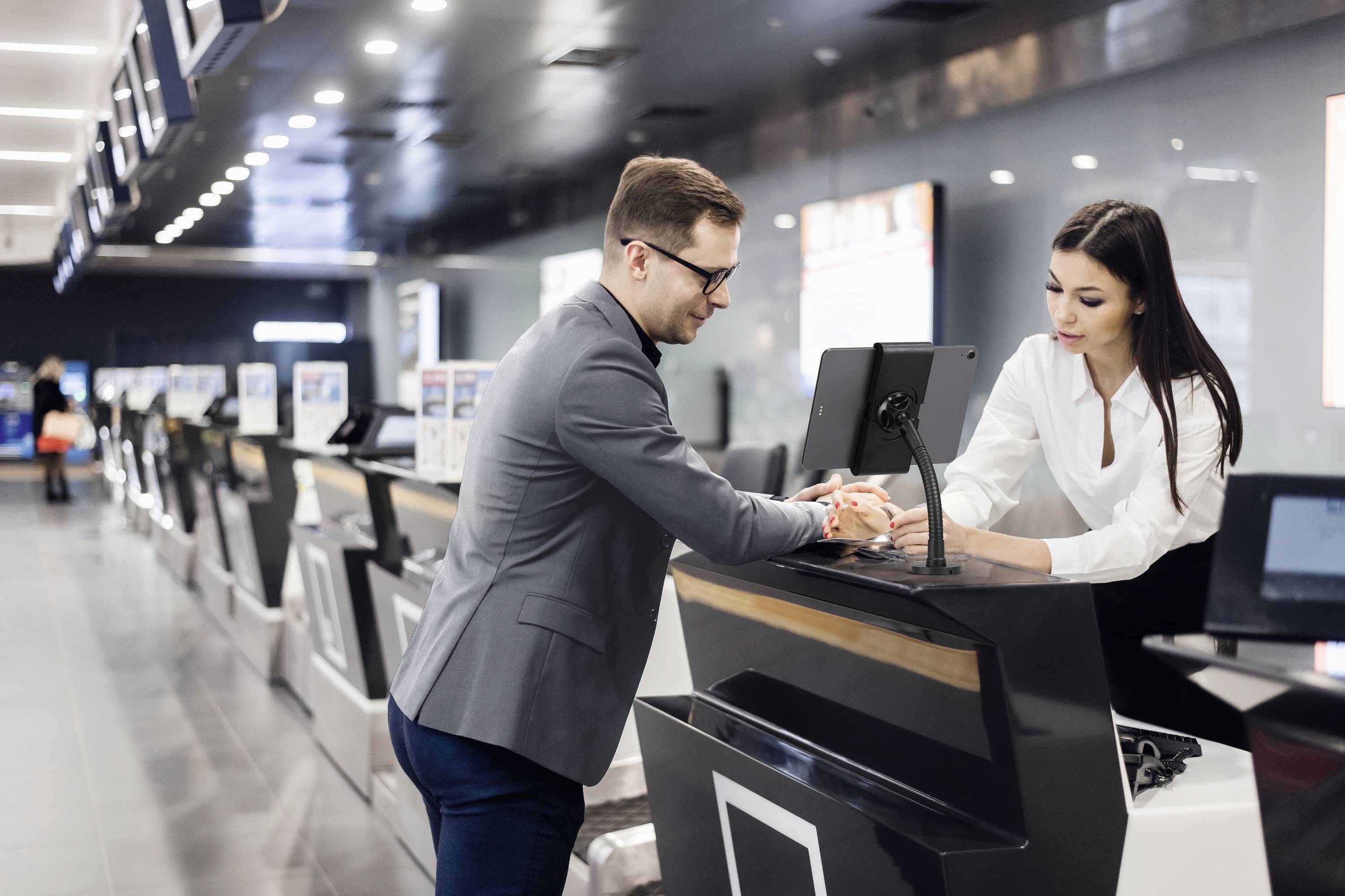 A man is speaking to a woman at a counter in a modern airport or office. The environment appears bustling and technological.