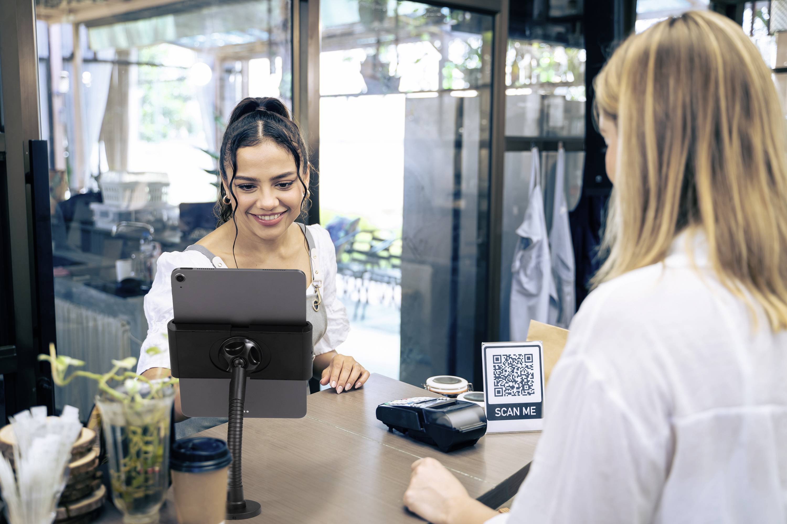 A woman behind a café counter smiles and helps a customer pay using a tablet. A QR code with 'Scan me' is on the counter.