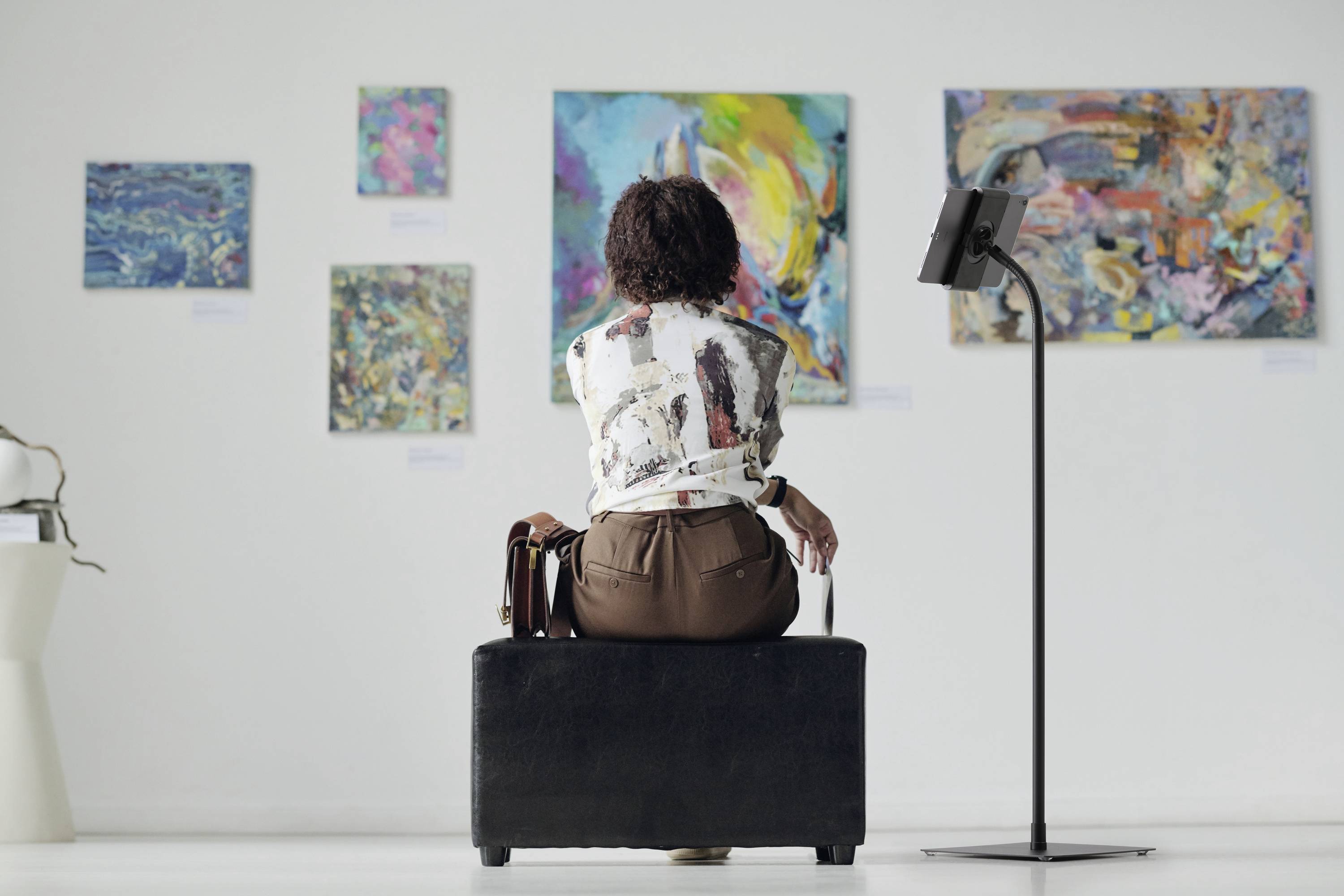 A person is sitting on a stool in an art gallery, looking at several abstract paintings hanging on a white wall.