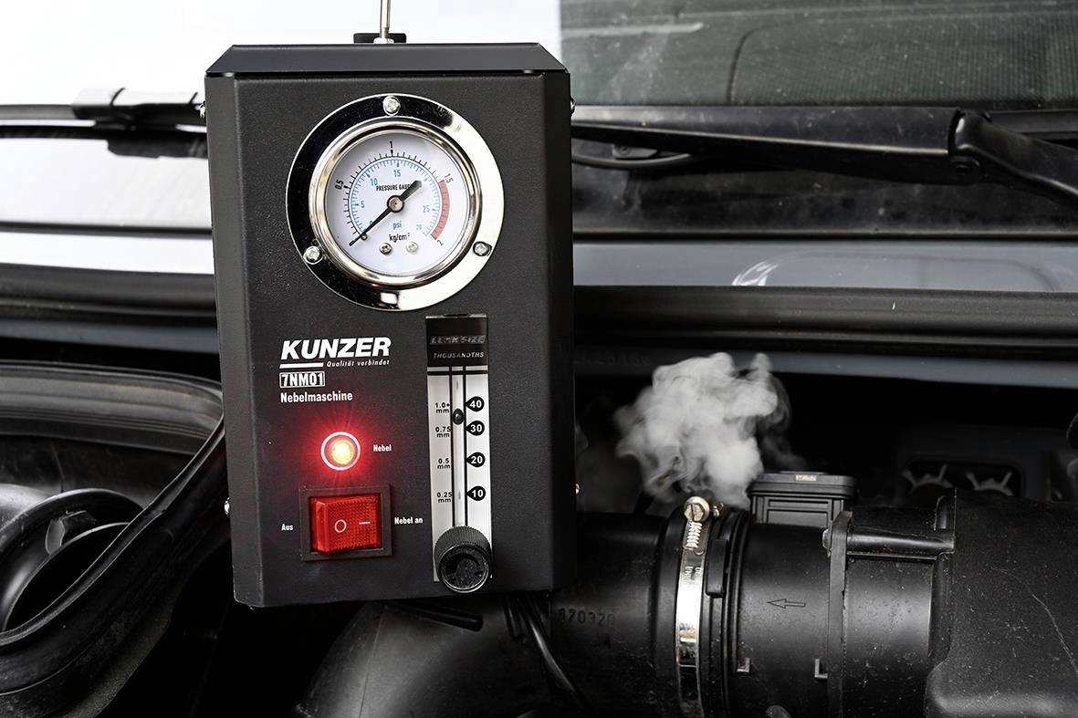 A diagnostic device for leak detection in a car engine. It shows a pressure gauge, LEDs, and smoke escaping from a hose.