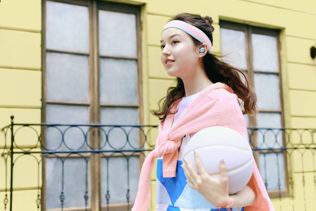 A young woman in sporty attire is holding a basketball and walking along in front of a building with a smile. She is wearing a headband.