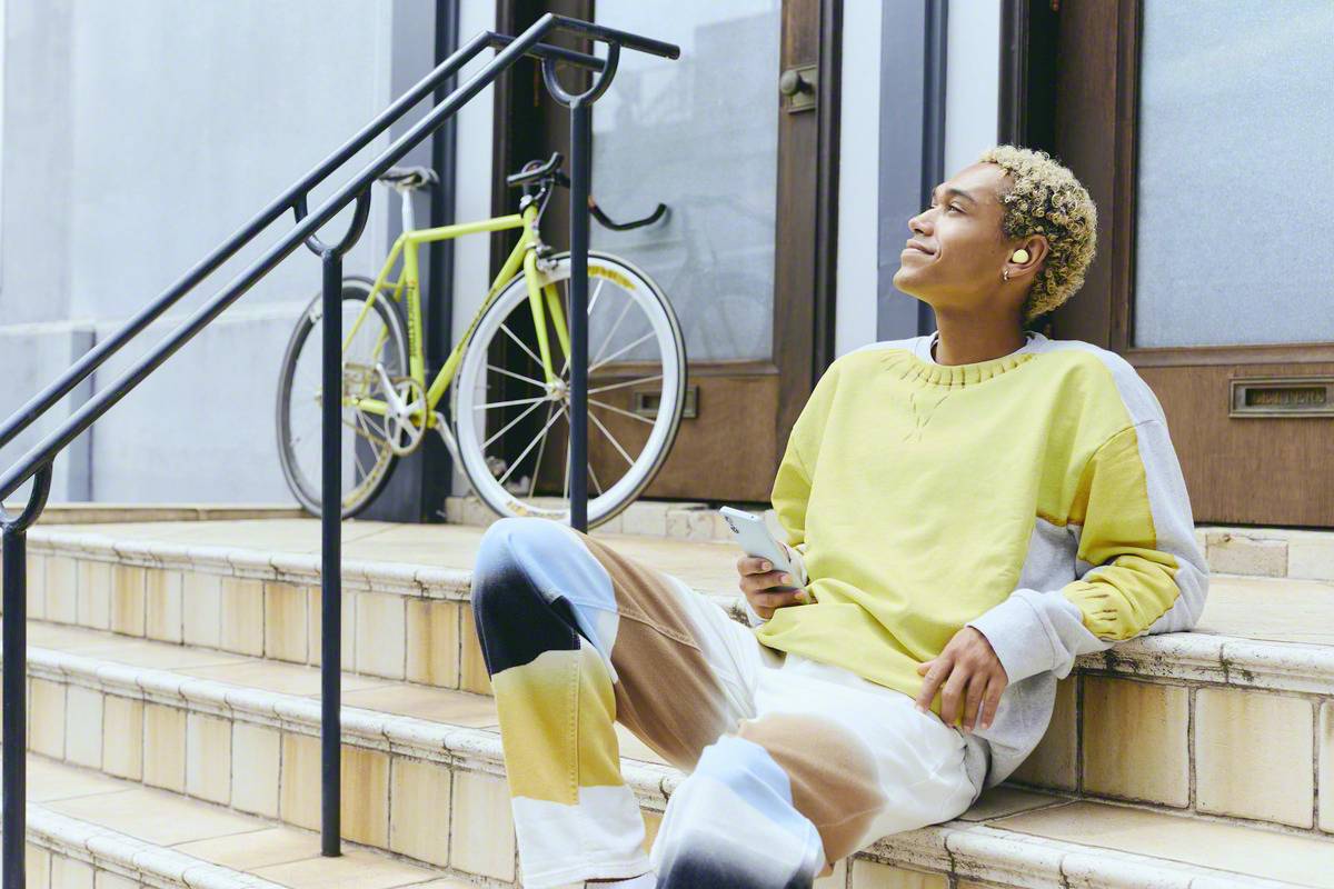 A person is sitting on a step, listening to music through earphones and holding a smartphone in their hand. A bicycle is standing in the background.