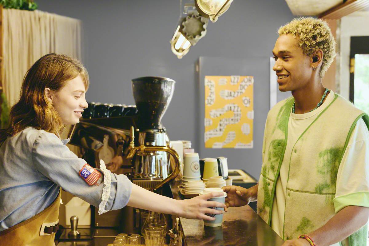 A barista passes a coffee across the counter to a customer in a cosy café. Both people smile warmly.