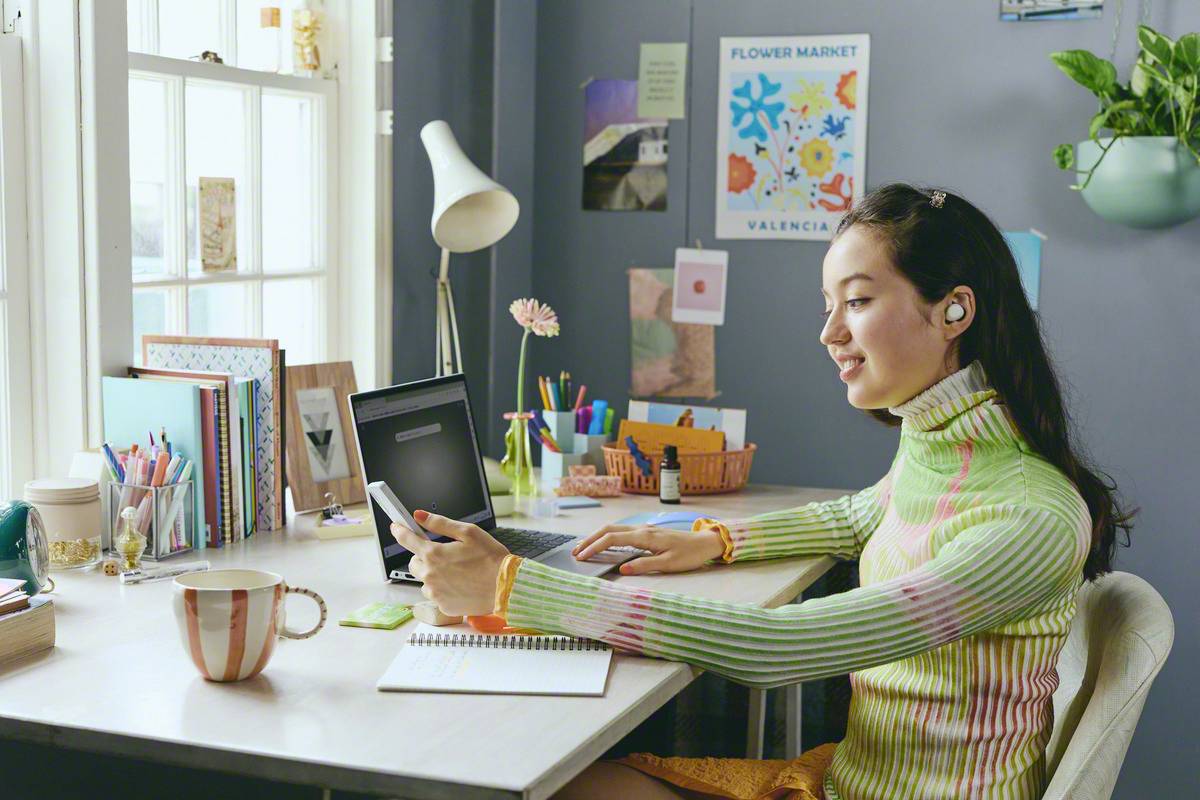 A smiling person is sitting at a desk, using a tablet and wearing wireless earbuds. The room is bright and creatively designed.