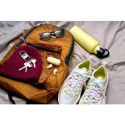 An orange rucksack is lying on a bed. Next to it are a yellow water bottle, trainers, glasses, a red headband, keys and earplugs.