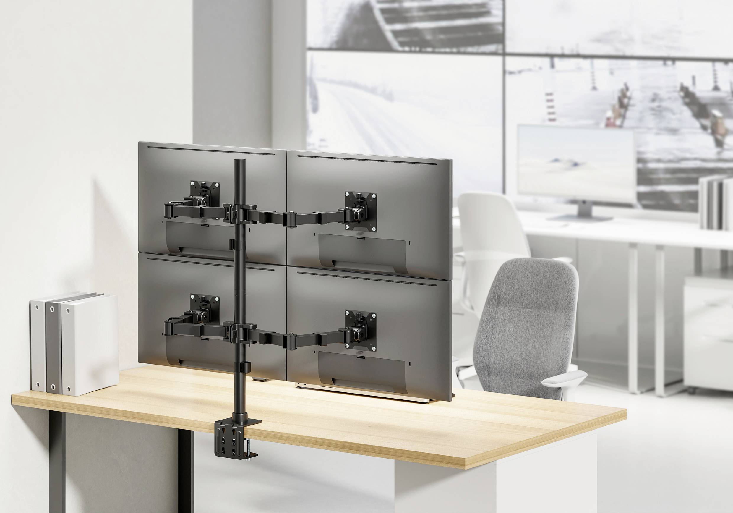 A desk with a monitor mounting system holding four screens above the desktop. In the background, a modern office.