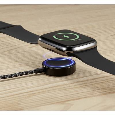 A smartwatch sits on a wooden table being wirelessly charged. A charging dock with a glowing ring indicates the charging process.