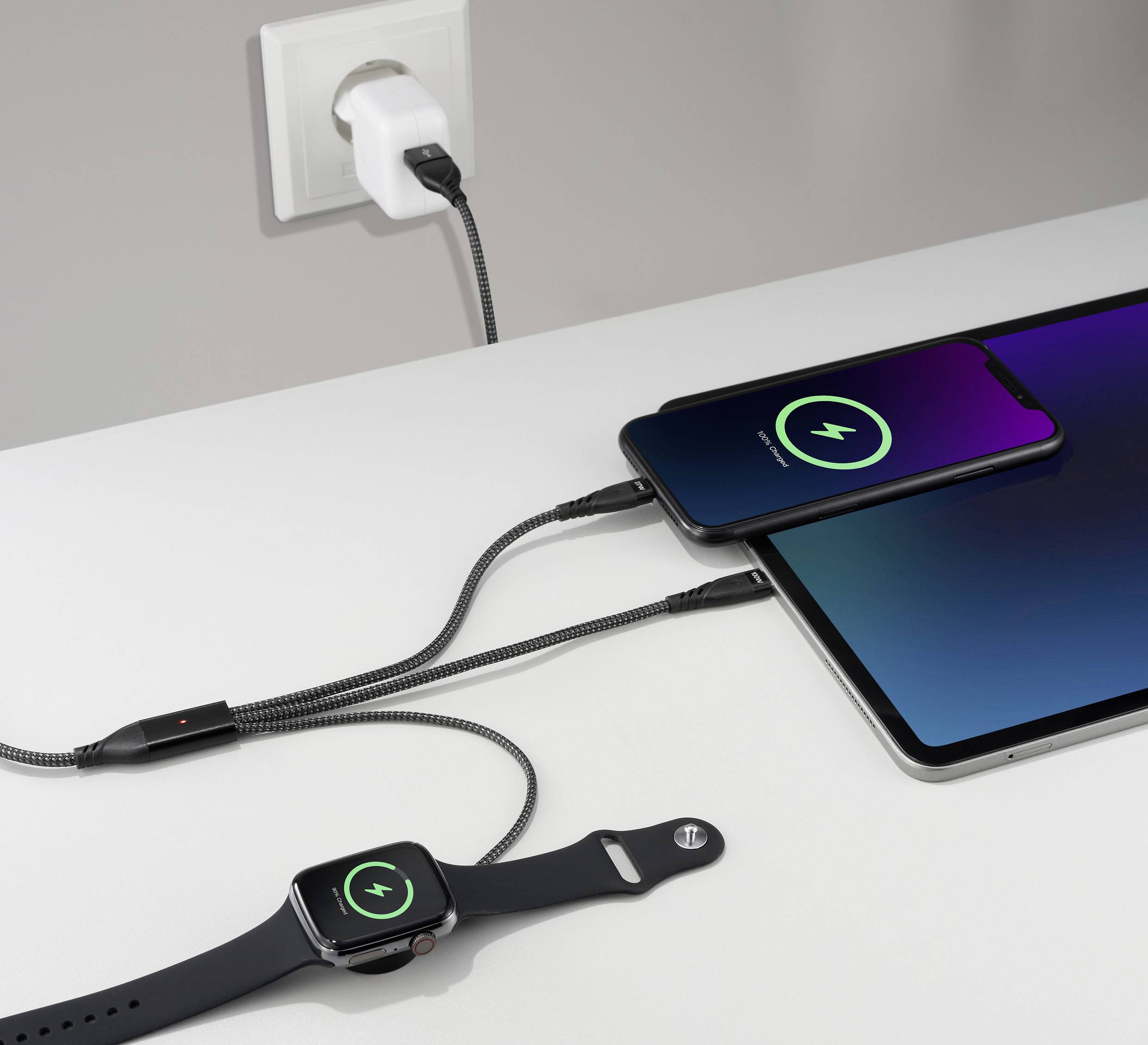 A smartphone, a tablet and a smartwatch are being charged simultaneously via a multi-charging cable on a table.
