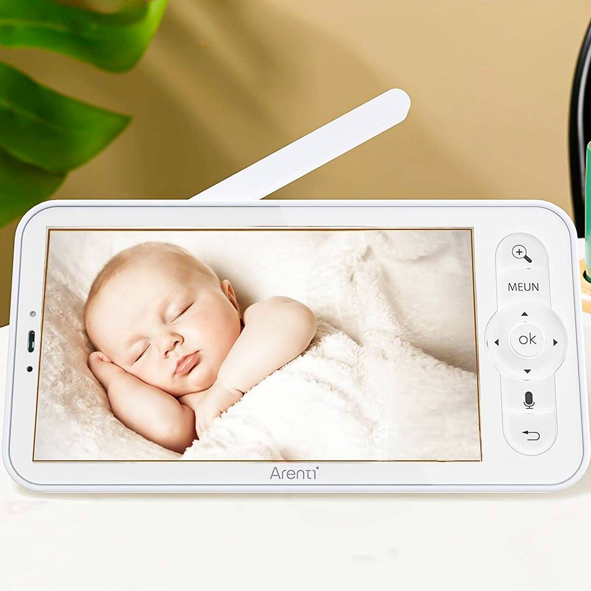A baby sleeps peacefully on a monitor with control buttons, surrounded by soft colours. A green leaf is visible in the background.