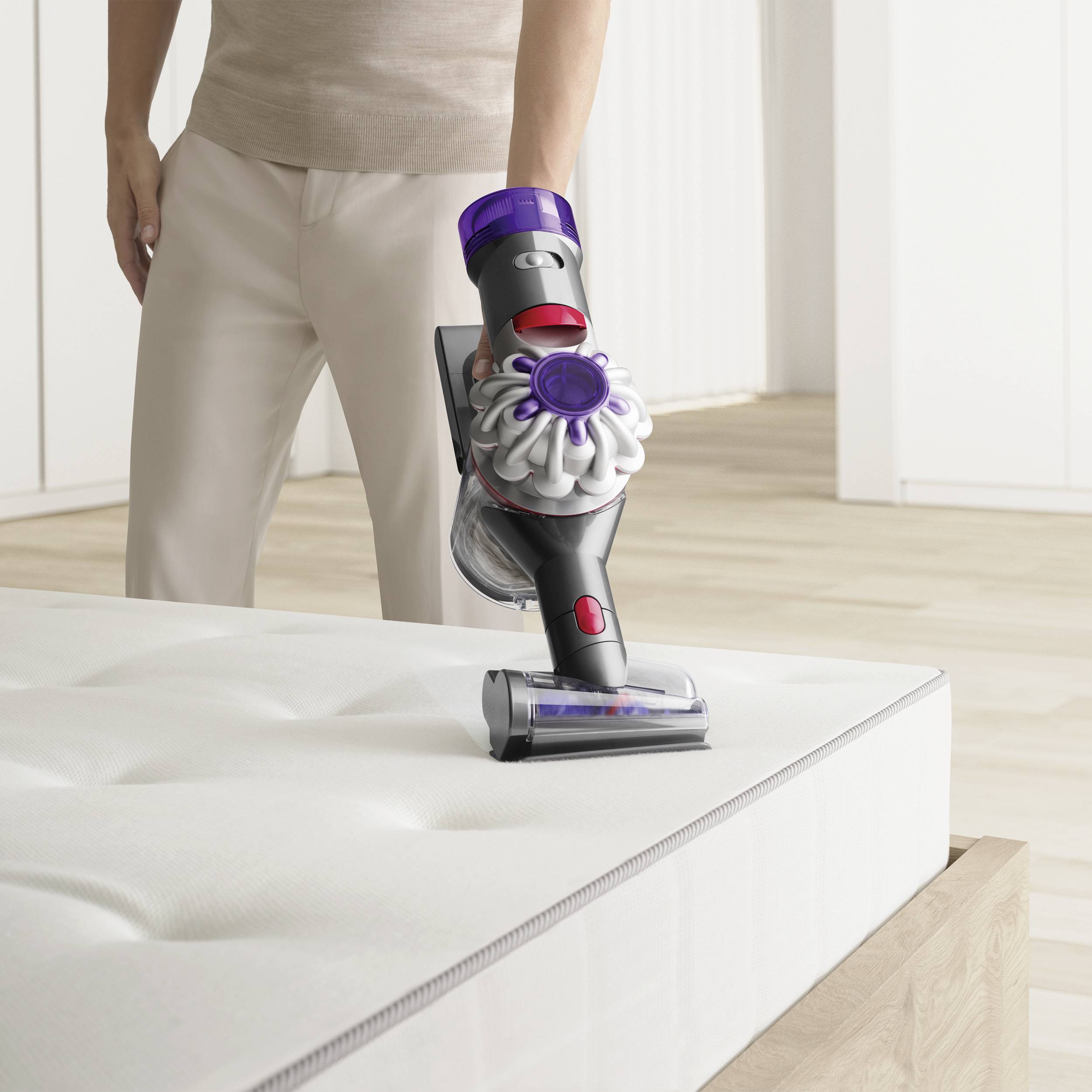A person is using a cordless vacuum cleaner to clean a mattress in a bright room. The vacuum cleaner has a modern design.