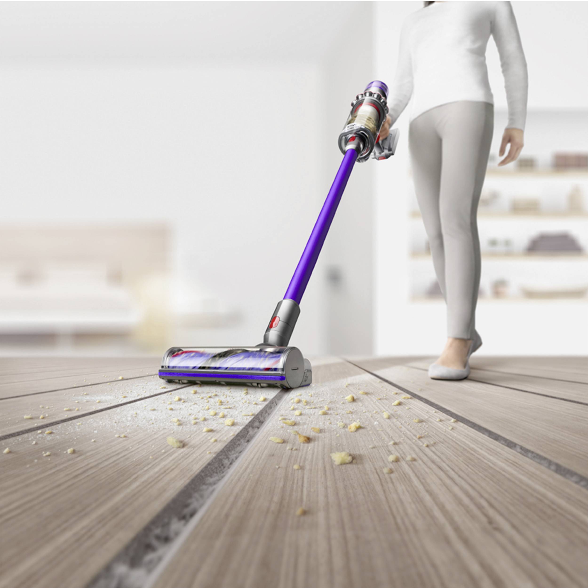 A person is using a cordless vacuum cleaner on a wooden floor, vacuuming up crumbs; a modern living room is visible in the background.