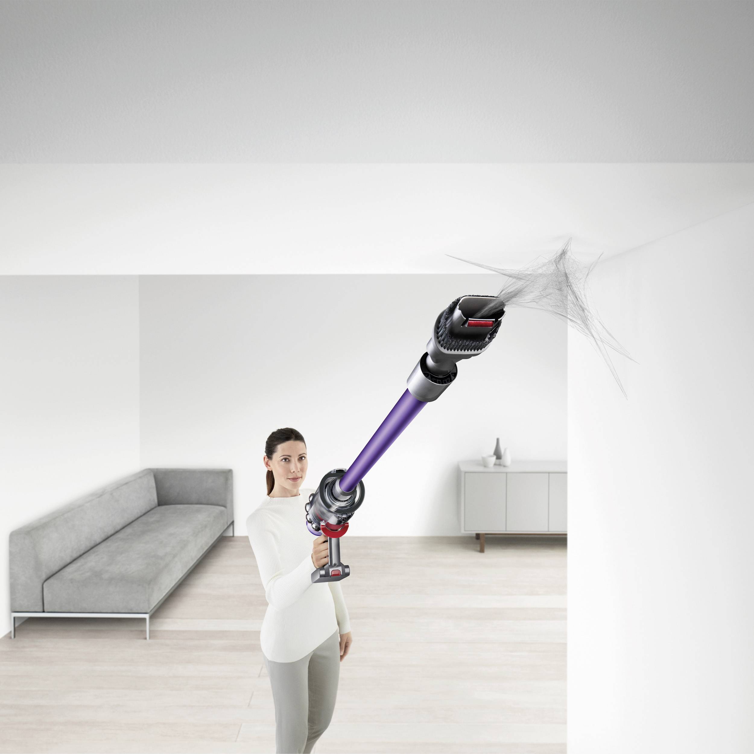 A person is using a cordless vacuum cleaner to remove cobwebs from the corner of a white ceiling in a modern, bright living room.