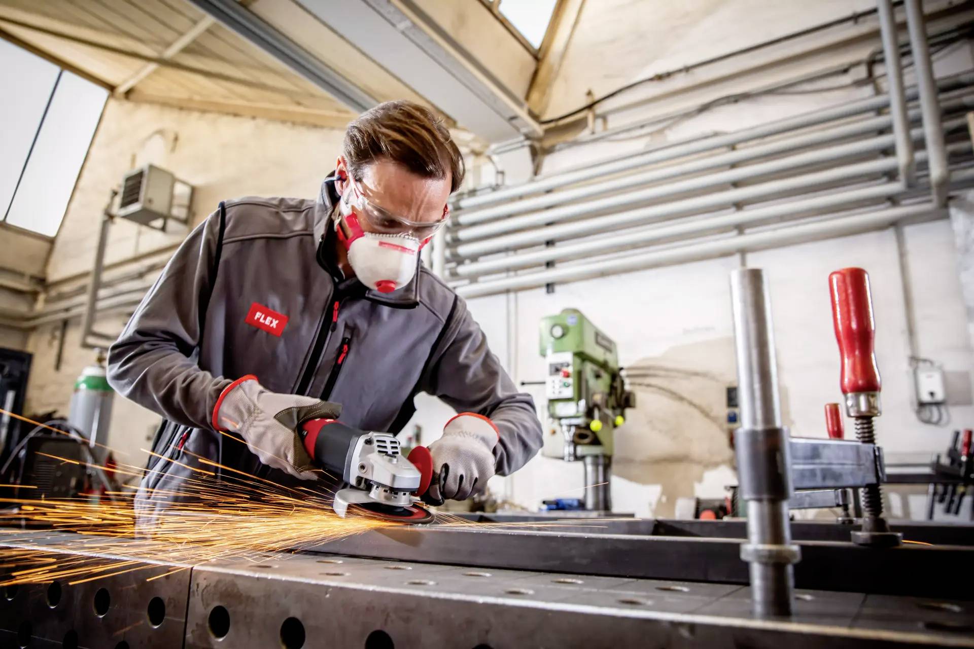 A worker wearing a protective mask and safety glasses grinds metal on a workbench, with sparks flying. Workshop environment with machinery in the background.