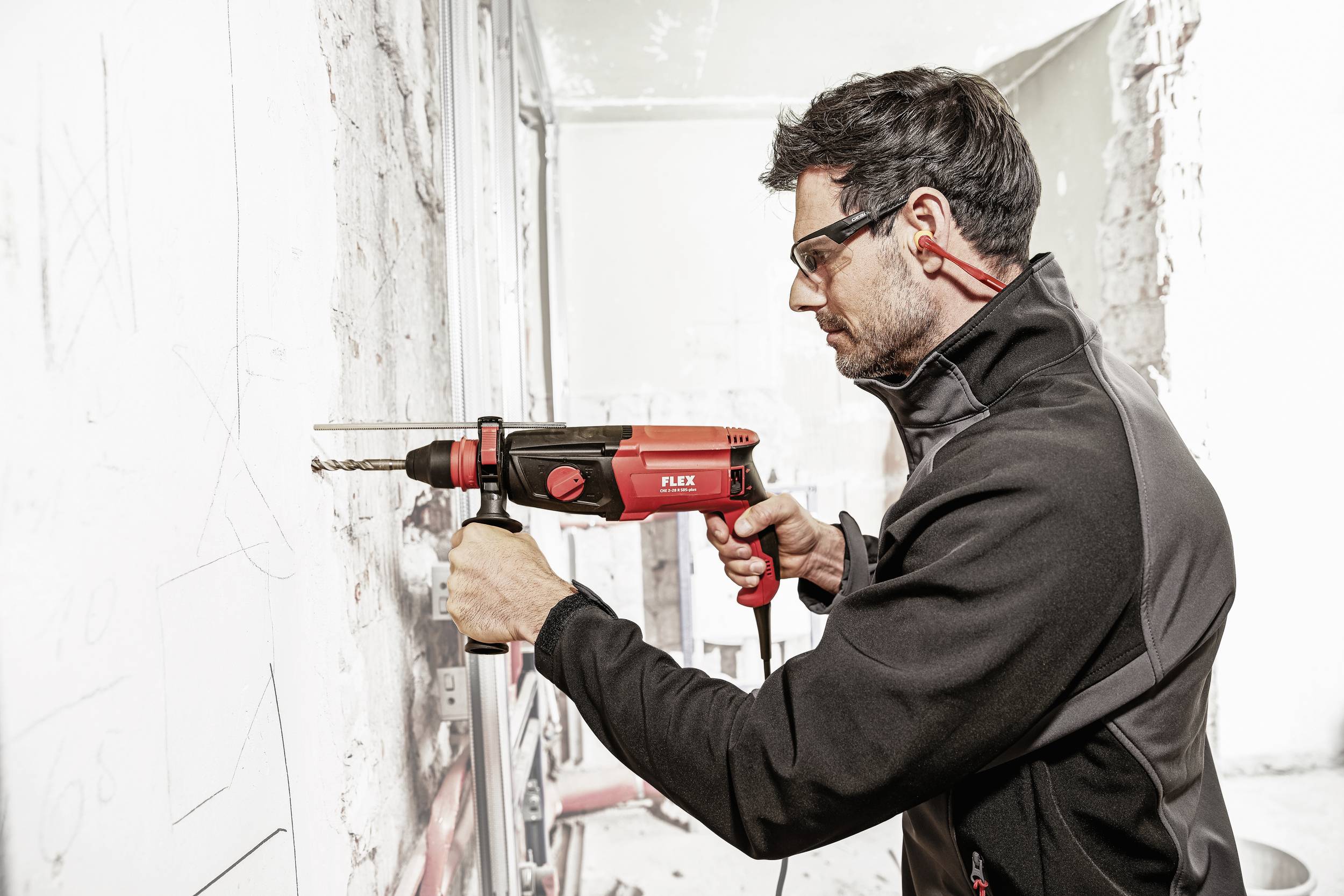 A man in workwear is using a red drill to bore into a white wall. Safety goggles and ear plugs are visible.