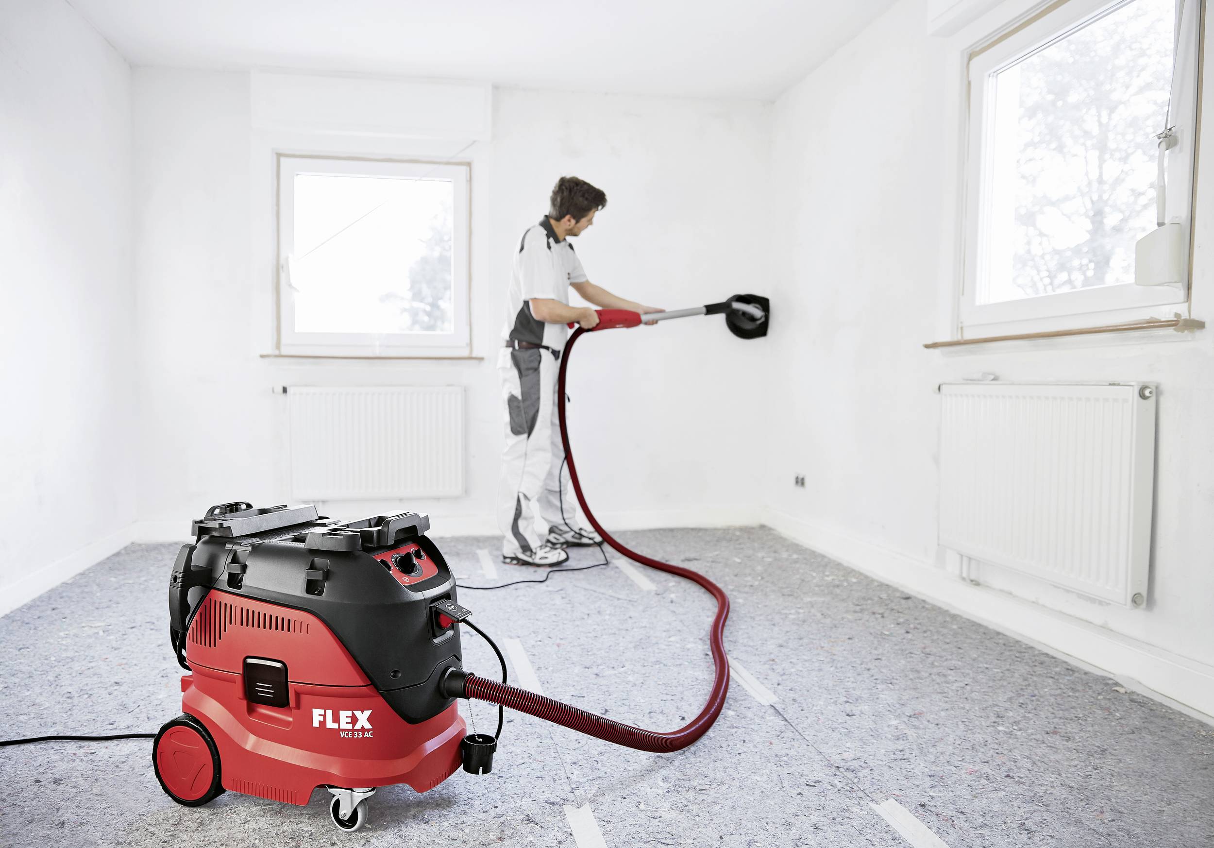 A tradesman is using a red industrial vacuum cleaner to sand a wall in an empty room. The floor is covered with a protective sheet.