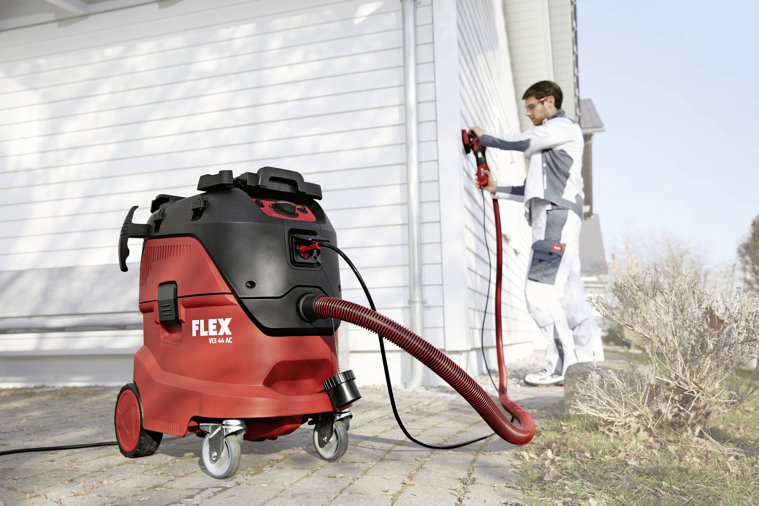 A red industrial vacuum cleaner by Flex stands in the foreground. In the background, a person is working on the exterior wall of a building.