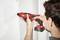 A person is using a red electric screwdriver to turn a screw into a white wall.