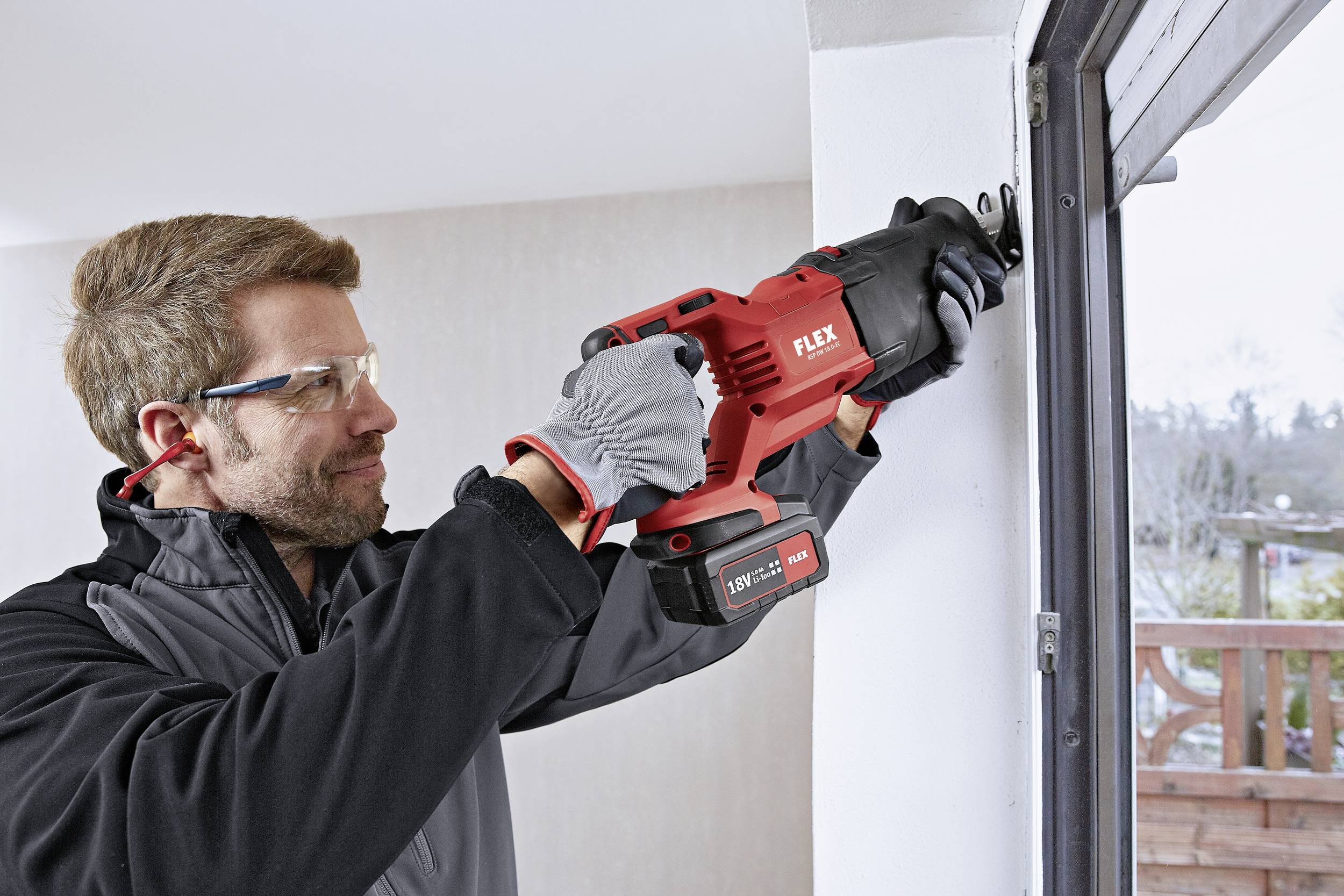 A man wearing safety glasses and gloves is using a red electric saw to work on a frame in a room with windows visible.