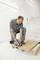 'A man in grey workwear is using a circular hand saw to cut a wooden board while kneeling on a ladder.'