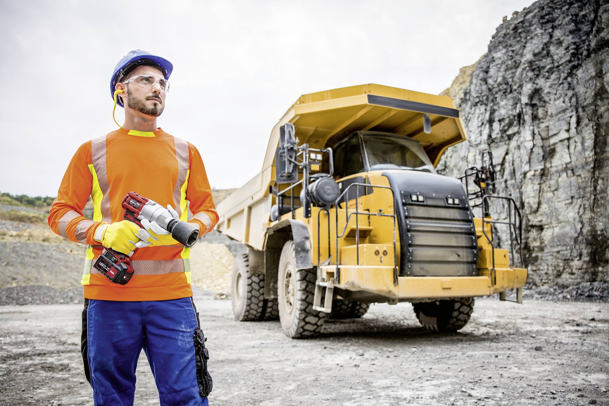 A worker in safety clothing stands in front of a large yellow dump truck in a quarry.