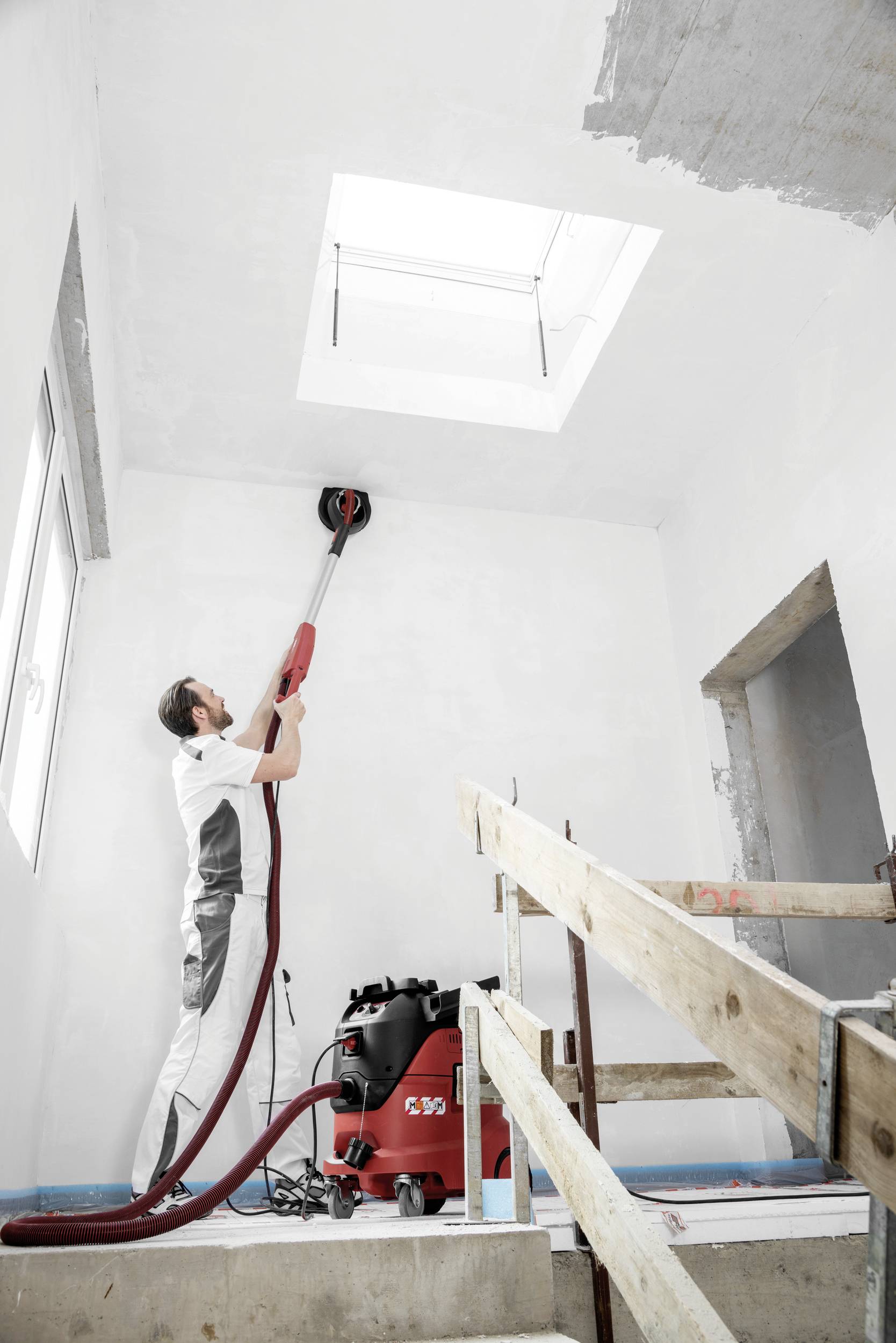 A person is grinding the ceiling of a construction site with a grinding machine. A red industrial vacuum cleaner is standing next to it.