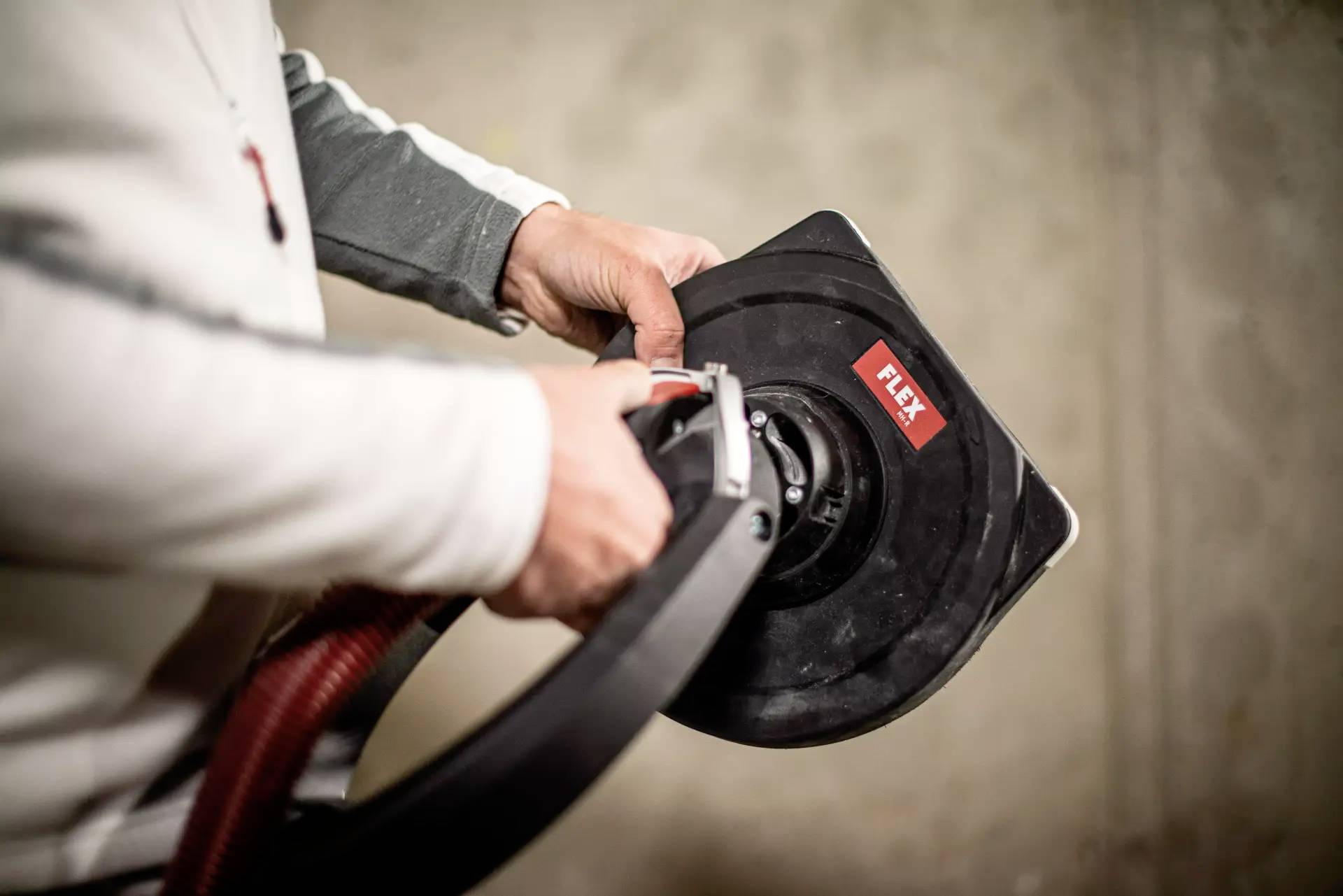 A person is holding an electric grinding tool with a red hose, of the Flex brand. The device is evidently being used in a workshop.