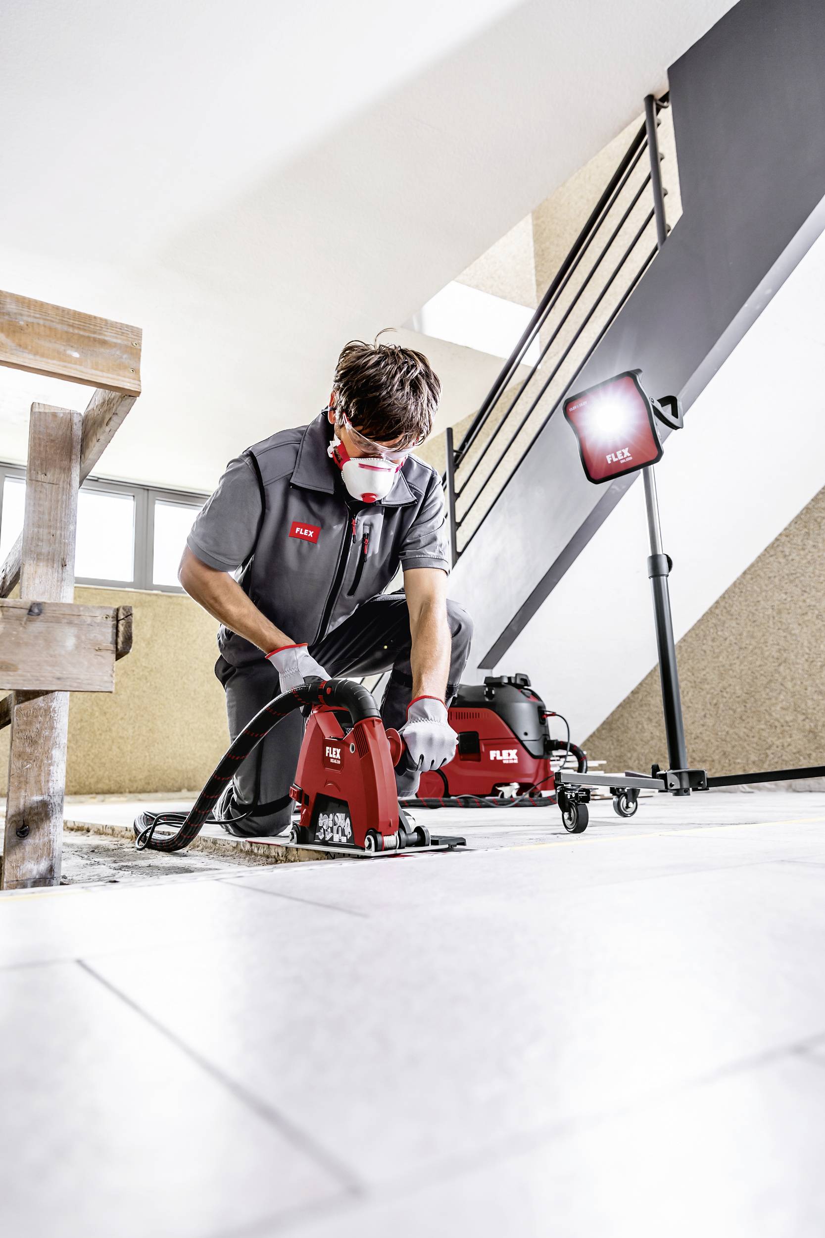 A tradesman in protective clothing is working with a joint cutter on a floor. A work light illuminates the scene.