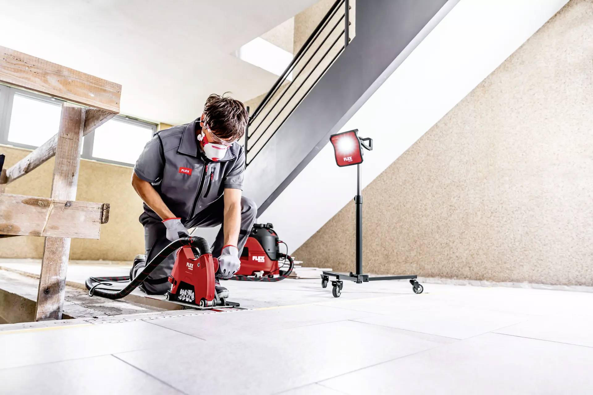 A worker in protective clothing is using a red construction device in a bright stairwell. A lamp is positioned next to him for illumination.