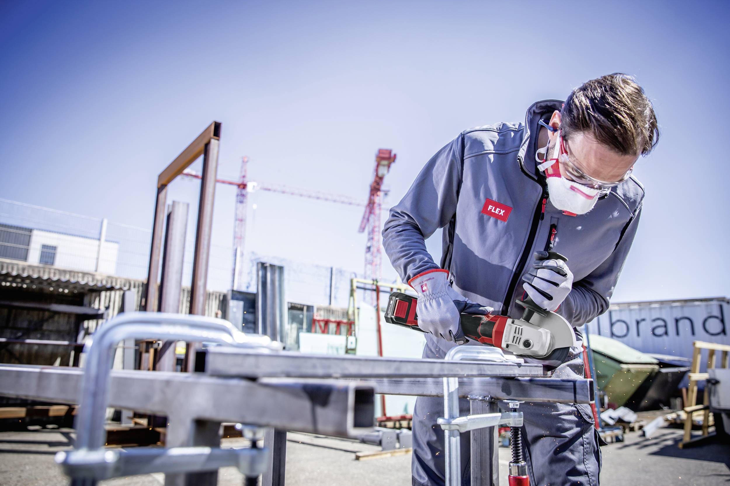 A man in protective clothing is working with an electric tool on a metal frame on an outdoor construction site under a clear sky.