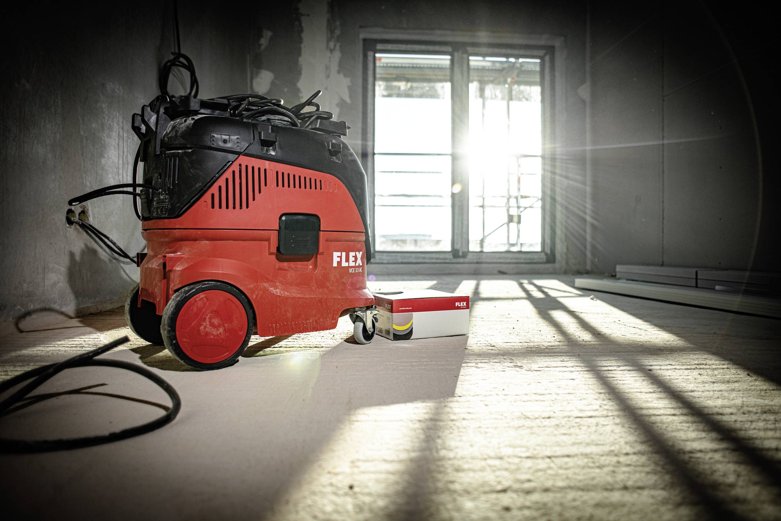 A red industrial vacuum cleaner sits on a raw concrete floor in an empty room, with sunlight streaming through a large window.