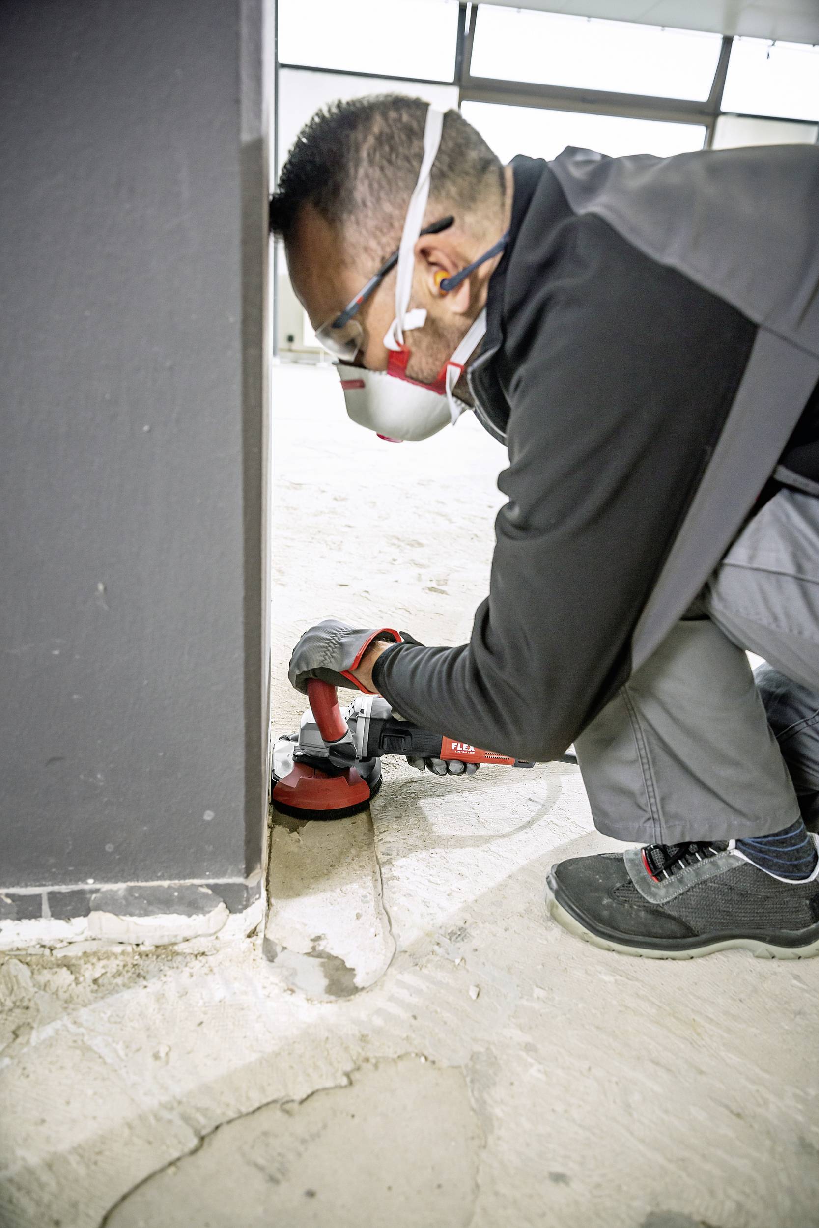 'Worker grinds floor next to concrete wall, wearing safety glasses, ear protection and dust mask. Focus on safety and precision.'