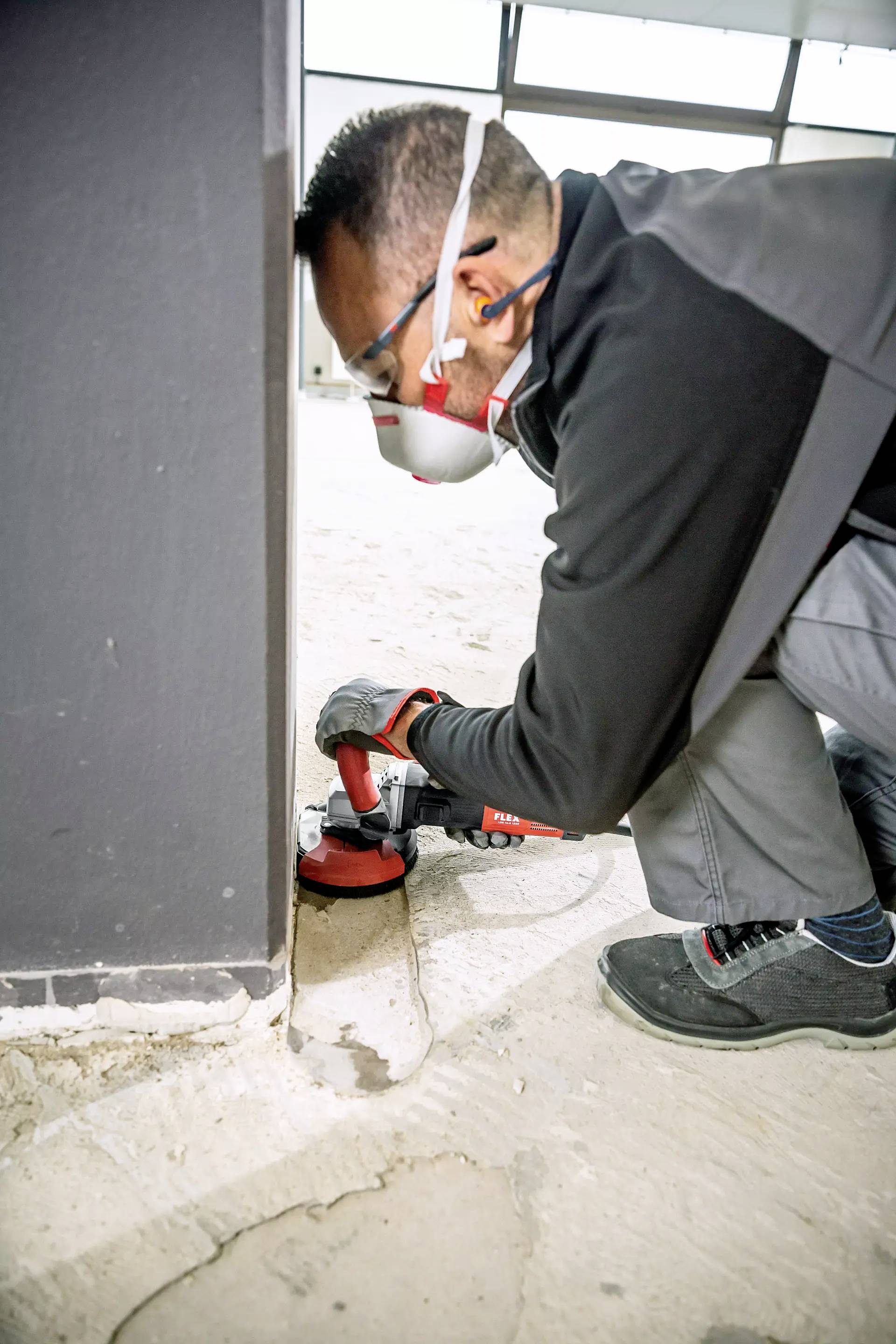 A worker is grinding the floor in an industrial building. He is wearing safety glasses, a respiratory mask and ear protection while using an electrical tool.