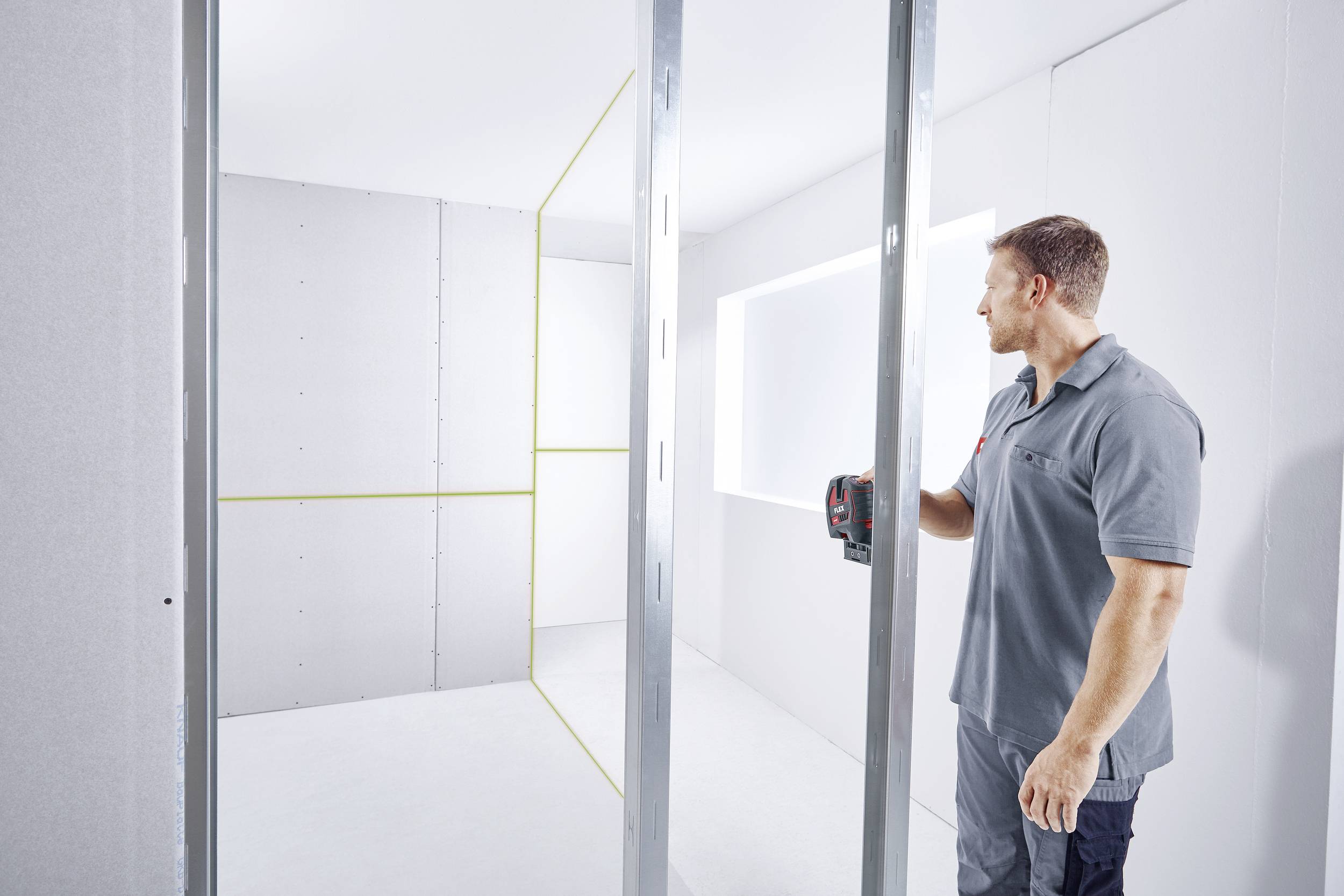 A man stands in a bright room with metal frames and plasterboard walls, holding a tool. The room has a large window and an incomplete wall structure.
