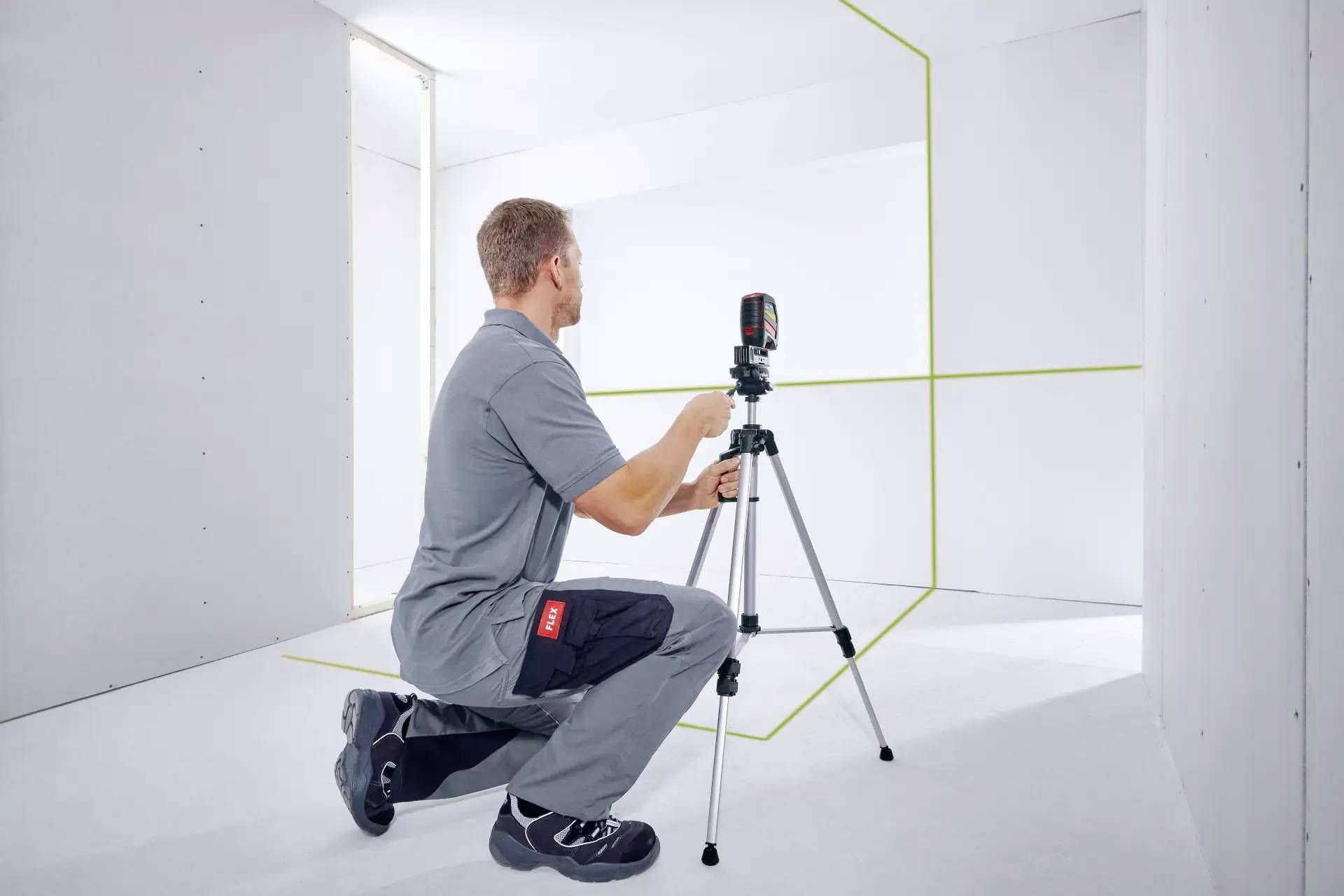 A man kneels in an empty room and adjusts a laser measuring device on a tripod. Green laser lines are visible.