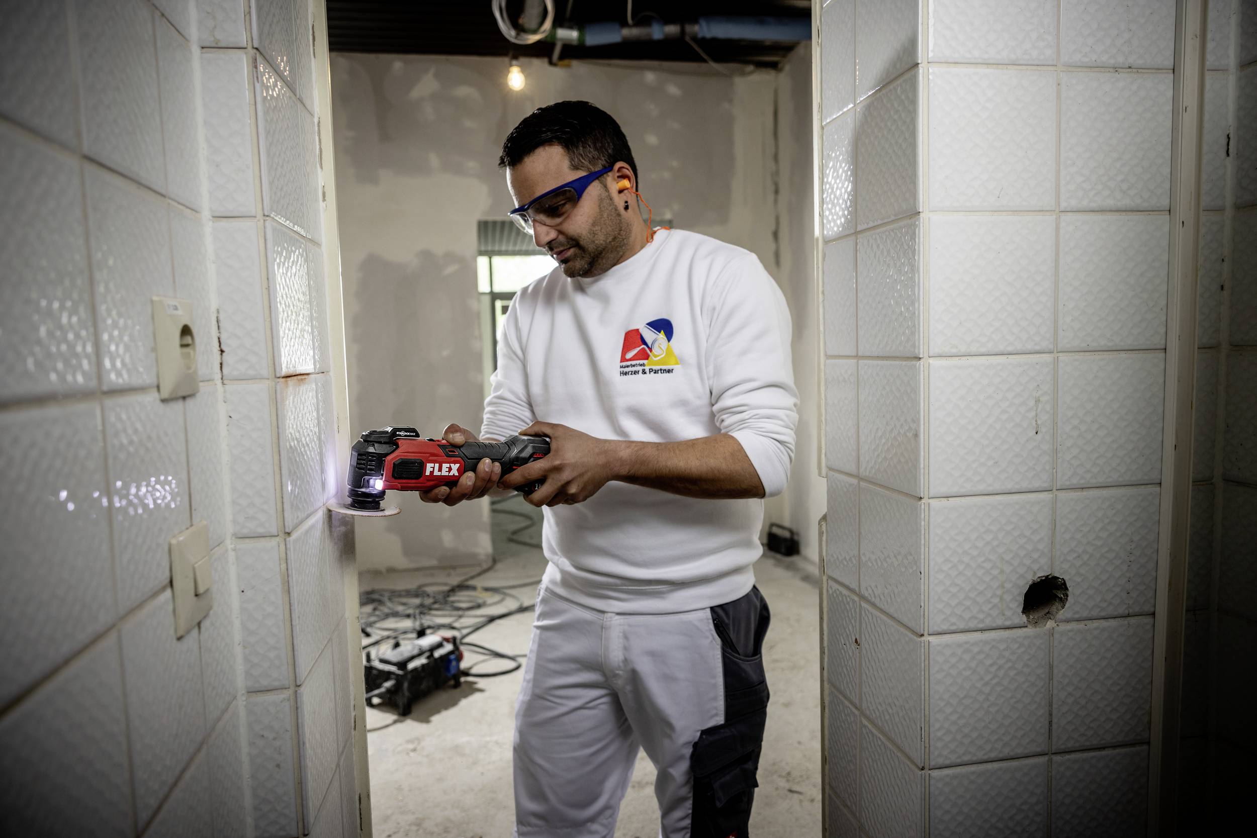 A man in work attire is using an electric tool on a construction site. He is wearing safety glasses and working with concentration on a wall.