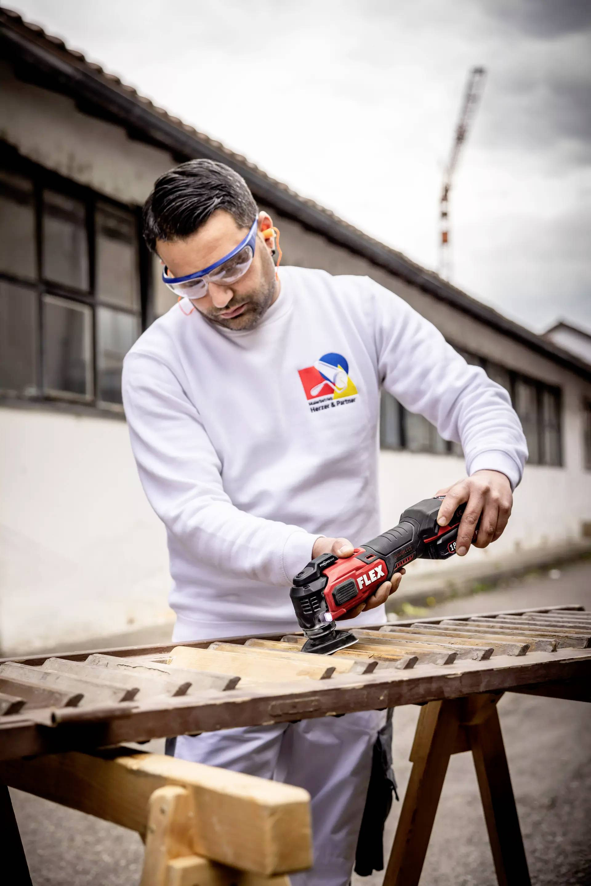 A man is sawing a piece of wood outside with an electric saw. He is wearing safety glasses and work clothing. Industrial buildings are visible in the background.
