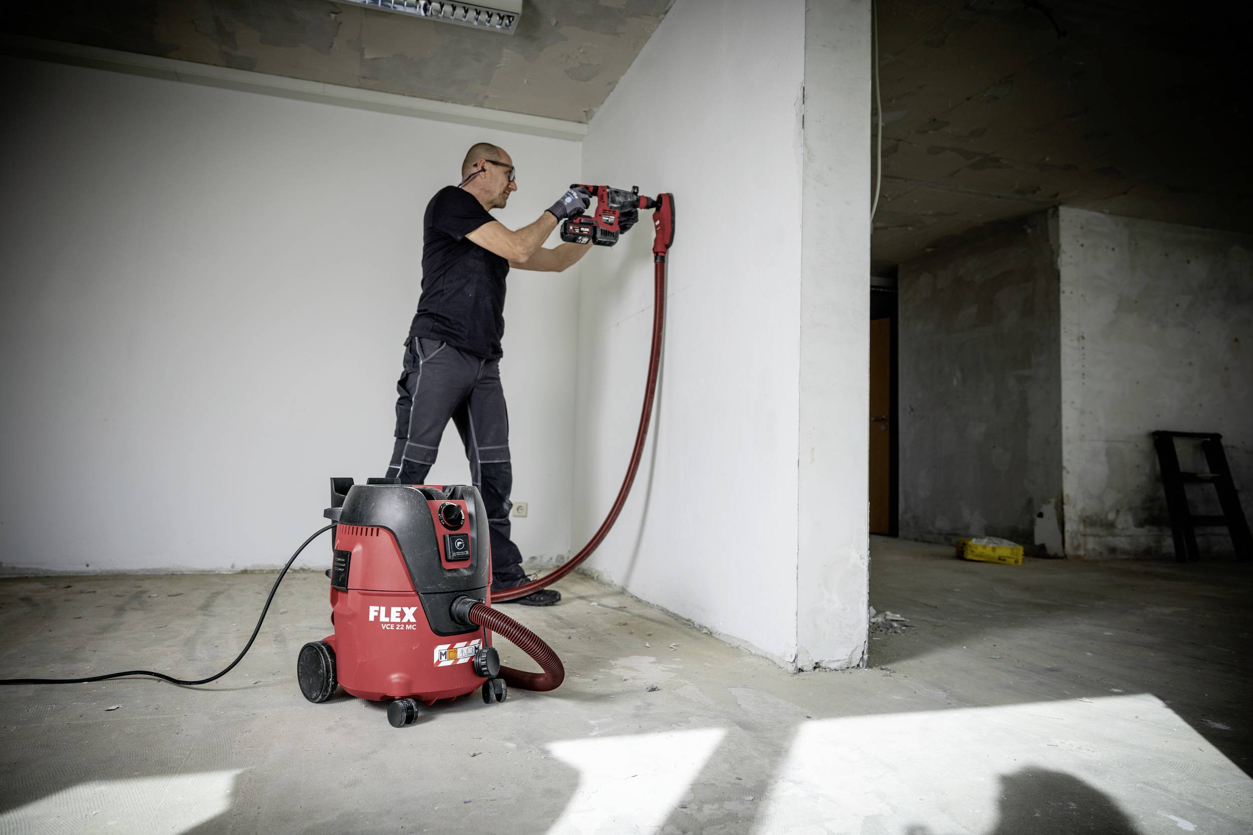 A person is drilling a hole in a white wall using an electric power tool in a room that resembles a building site. A red vacuum cleaner is standing nearby.