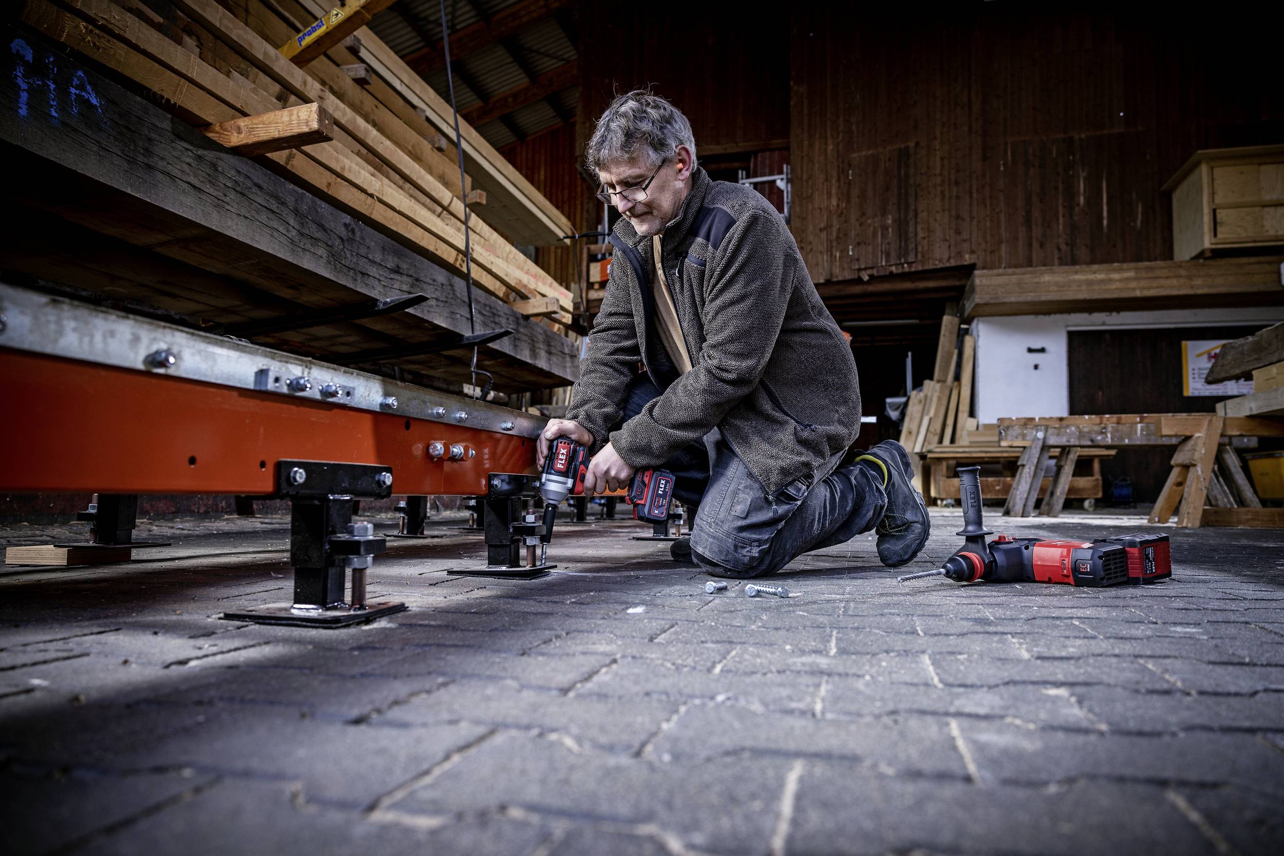 A man is kneeling on the floor in a workshop and working with electrical tools on a metal construction.