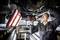 A mechanic is working underneath a raised vehicle, holding an electric tool, with a lamp illuminating the workspace.